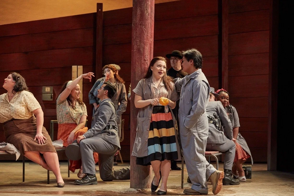 Actors on stage in a theatrical production, engaged in a dramatic scene, with several other actors seated and standing in the background.