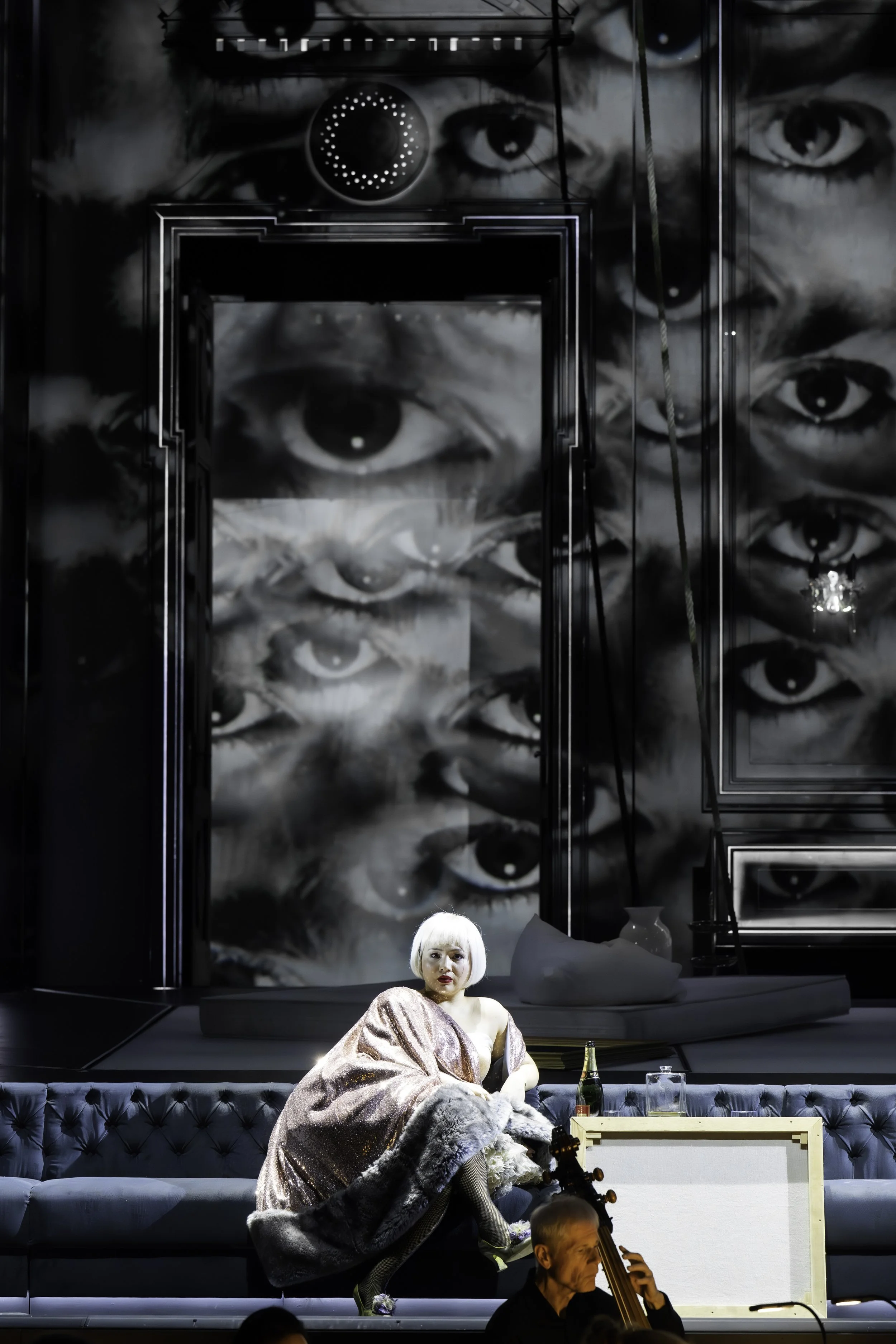 A woman with platinum blonde hair sitting on a stage in front of a black and white mural of multiple eyes. She wears a shiny, metallic dress and a faux fur coat. There is a cello player in front of her and a bottle, a glass, and a guitar amplifier on