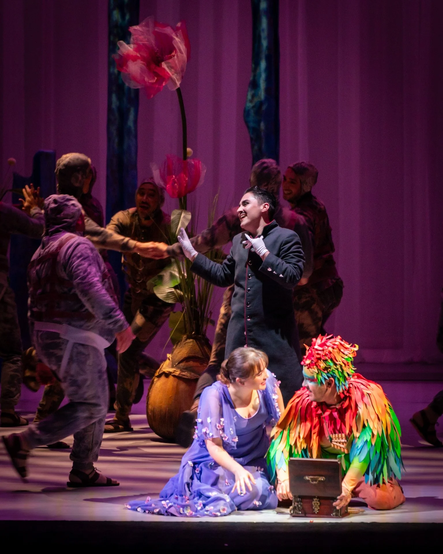 Actors on stage performing in a theatrical play with colorful costumes, large flower prop, and a treasure chest, under vibrant stage lighting.