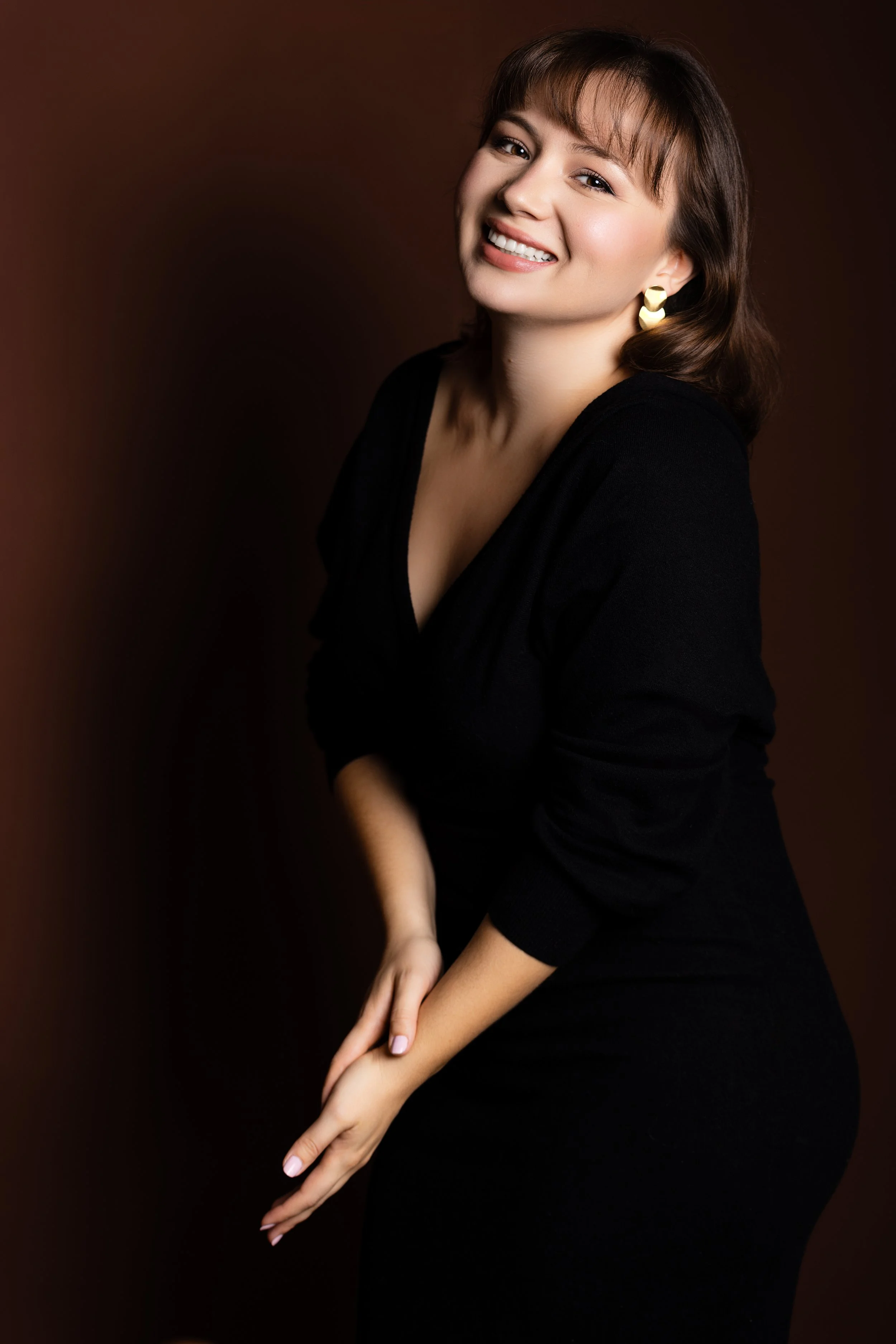 A woman with brown hair smiling, wearing gold earrings and a black top, against a dark brown background.