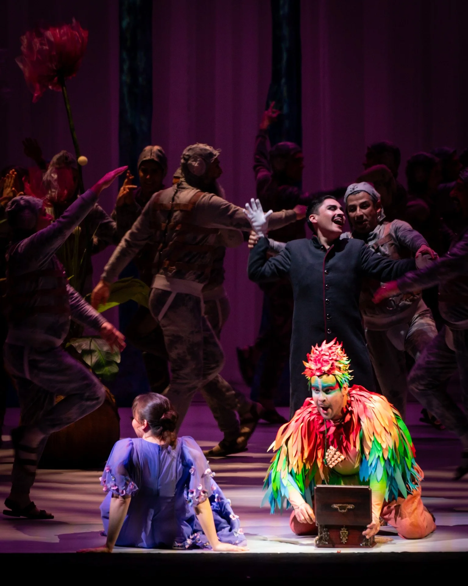 The image shows a theatrical stage scene with performers in colorful costumes. Two characters in the foreground are on the ground, one in a vibrant feathered costume and the other in a purple dress. The background features actors dressed in military-