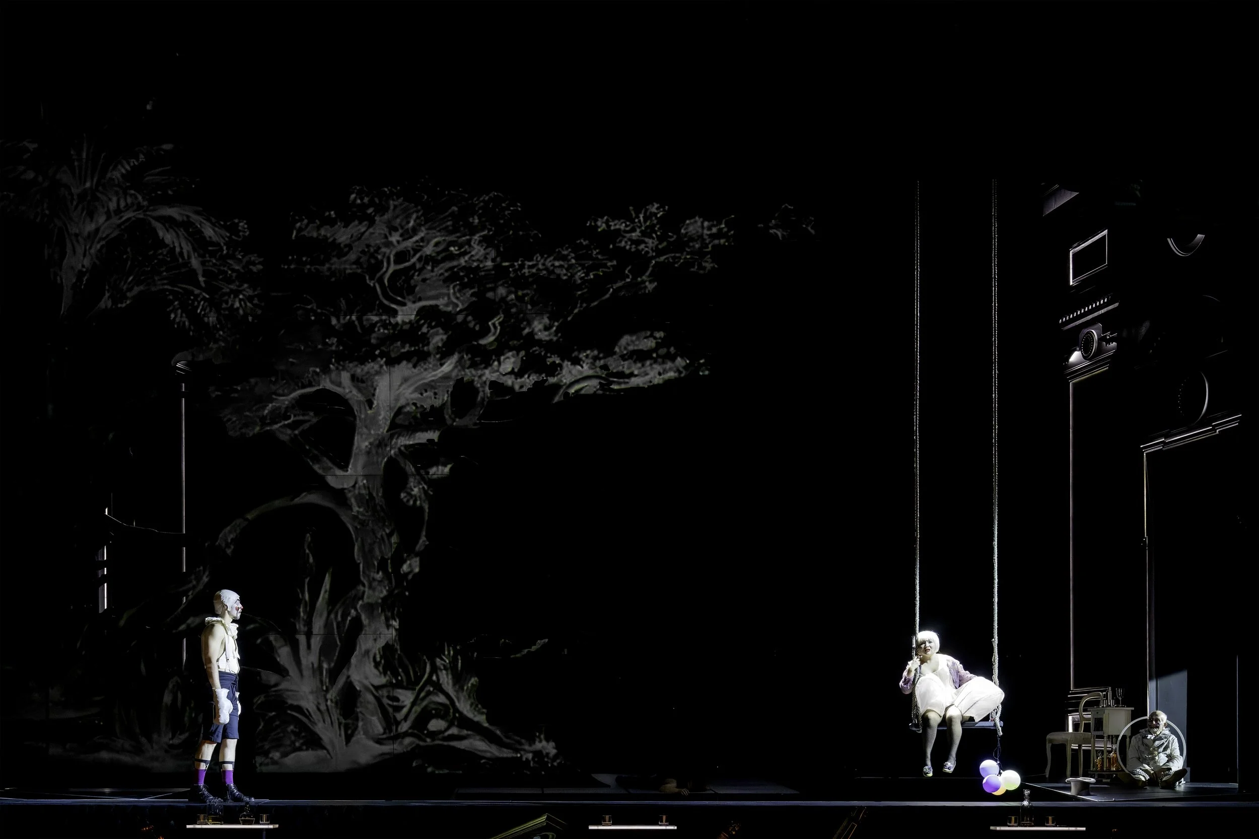 A theatrical stage scene with mannequins and mannequins wearing costumes, set against a dark background with abstract tree-like designs.