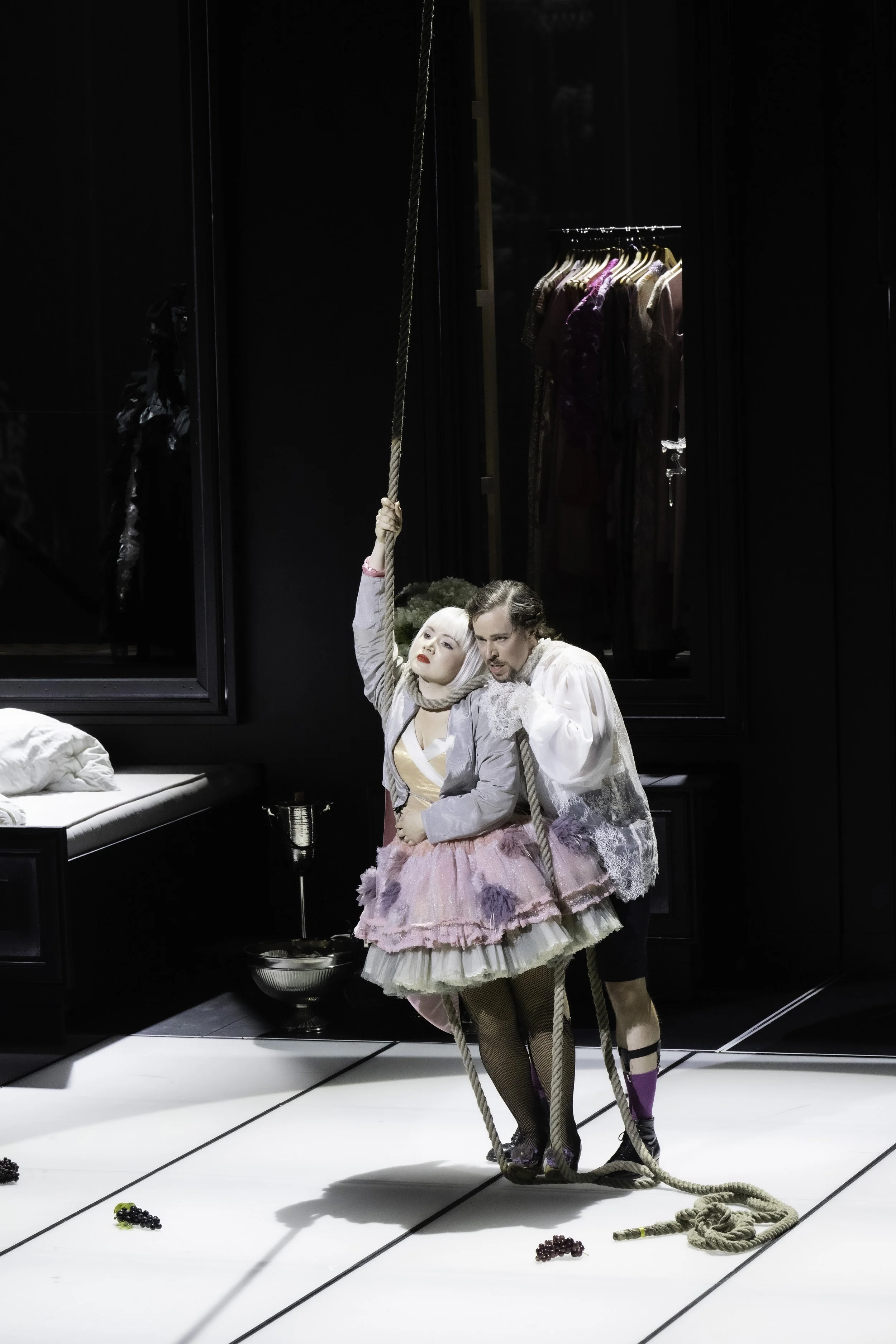 Two theater actors in period costumes performing a scene with a rope prop in a modern minimalist set. Woman in a pink petticoat and gray jacket, man in a white lace shirt and dark shorts, with beads and a candlestick on the floor.