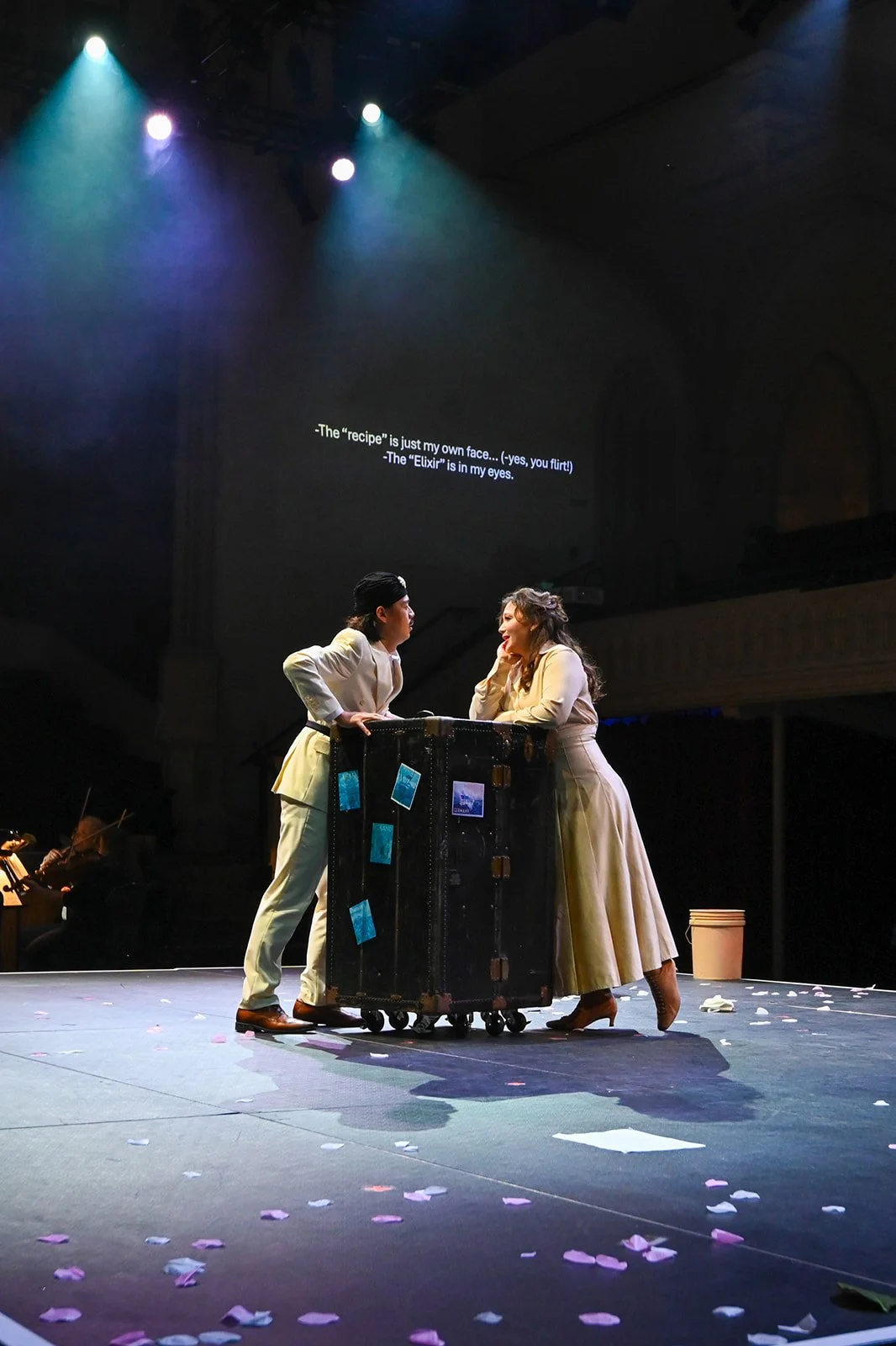 Two actors in vintage clothing on stage, leaning on a trunk, engaged in conversation. The stage is decorated with scattered confetti, and there is a dark background with spotlights shining down. Text is projected on the wall behind them.