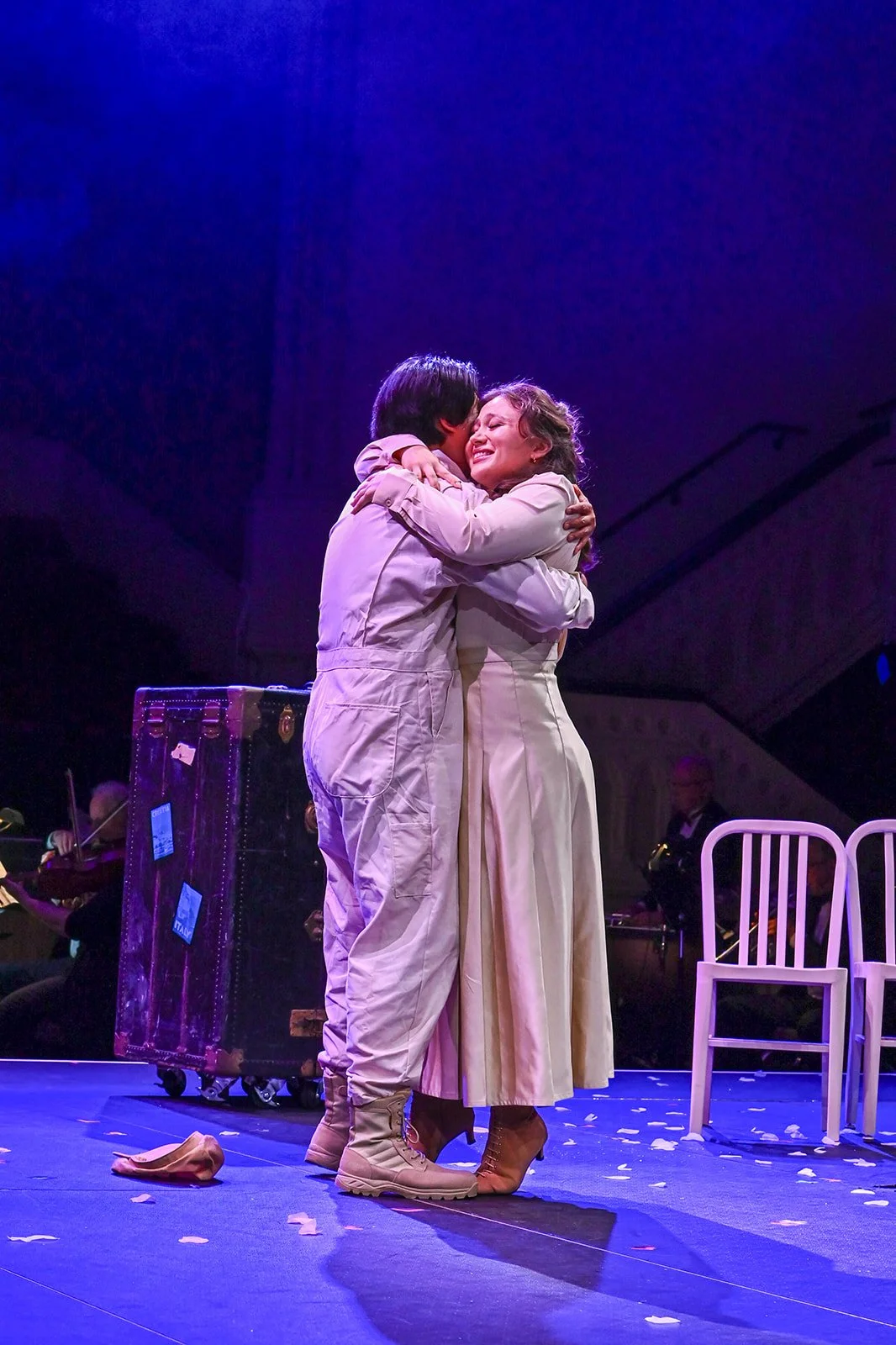 Two actors, one dressed as a soldier and the other in a white dress, embracing and smiling on stage during a performance, with violins and chairs in the background and scattered paper on the floor.