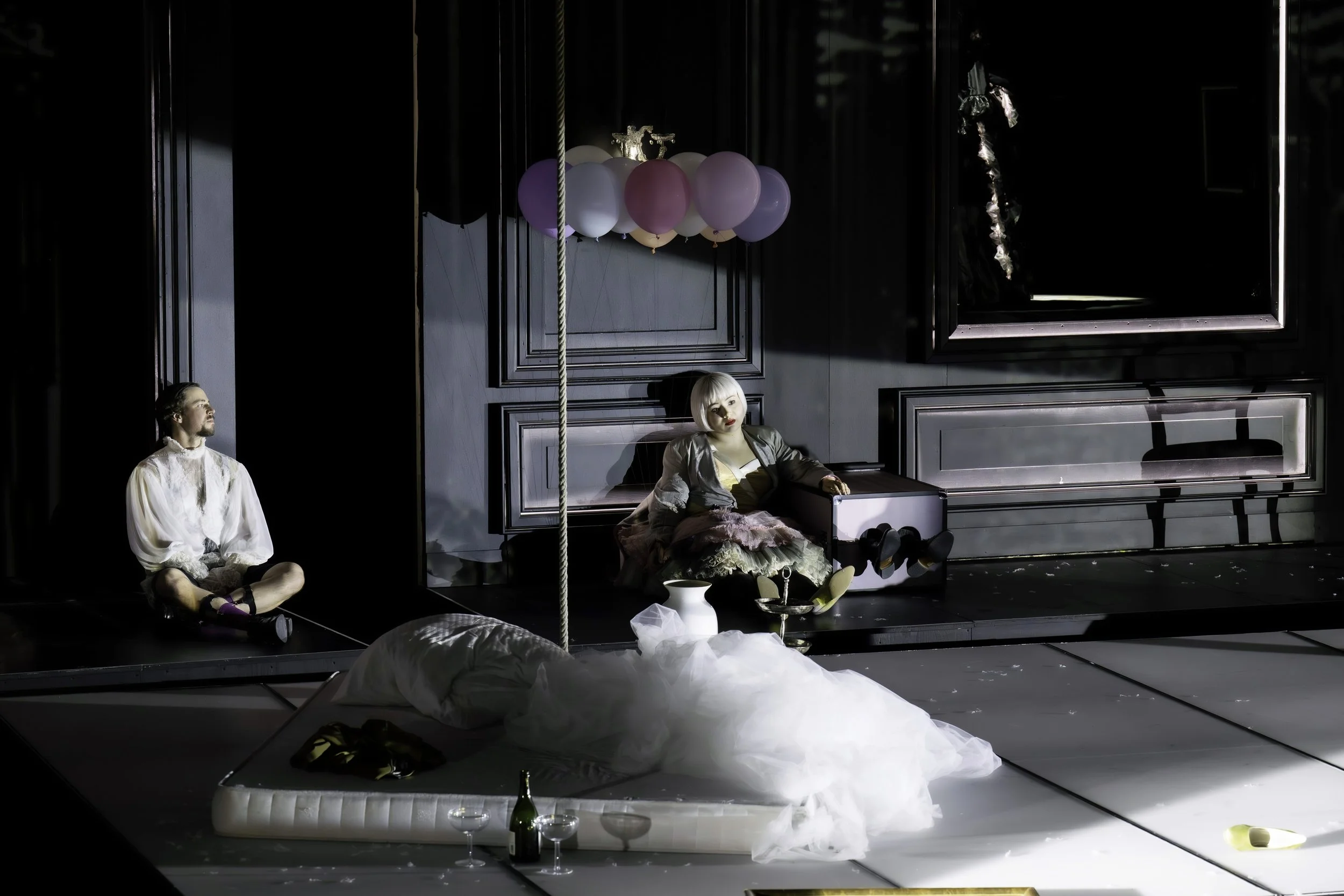 The scene appears to be a stage set with two actors seated on the floor in front of a dark, paneled wall. A cluster of balloons in pastel colors hangs above them. One actor, a man, sits cross-legged on the left, looking to his right. The other actor,