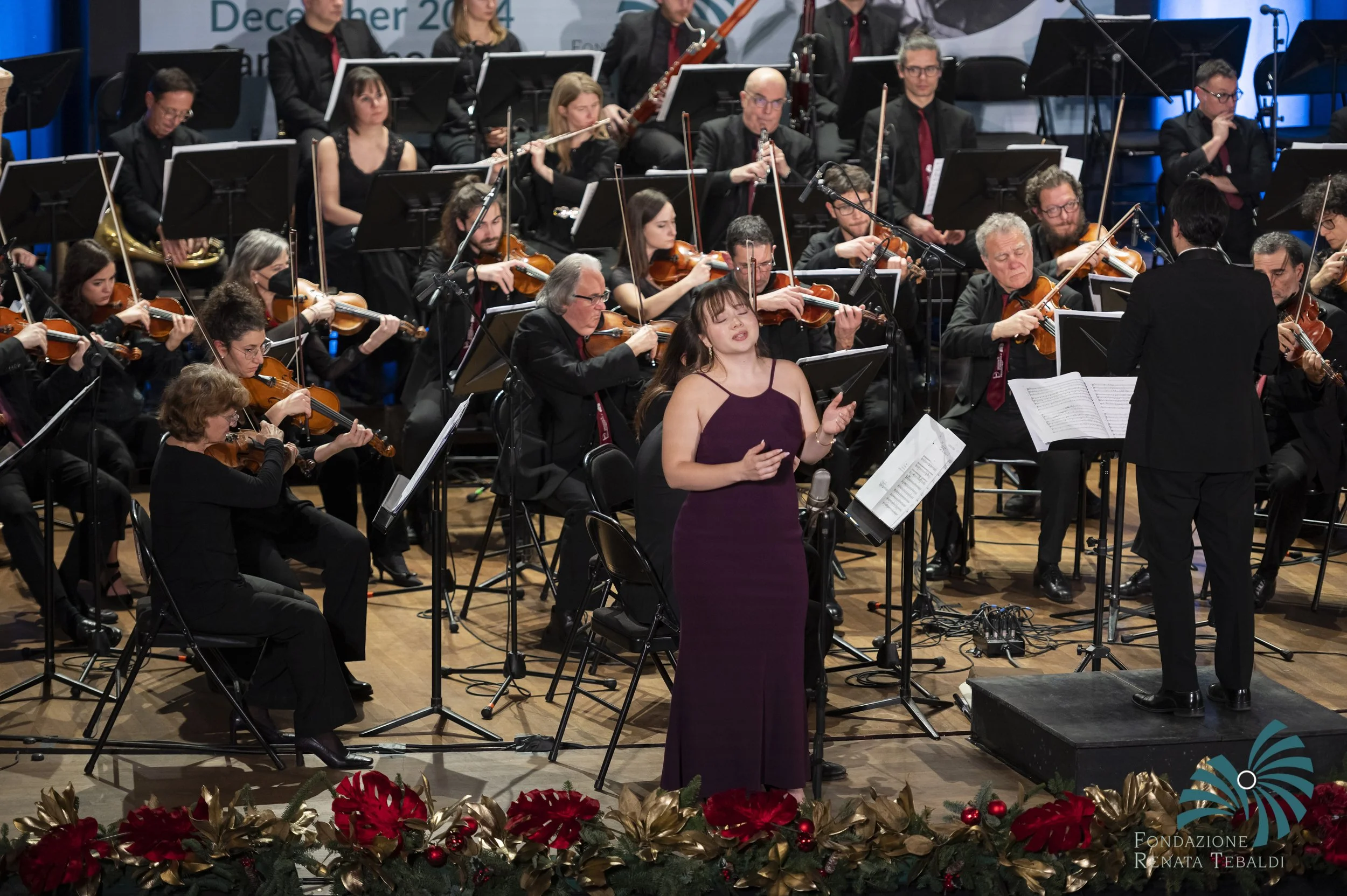 An orchestra performing on stage with a woman singing in front. The musicians are playing violins, cellos, and wind instruments, under the conductor. The stage is decorated with red poinsettias and festive decorations.