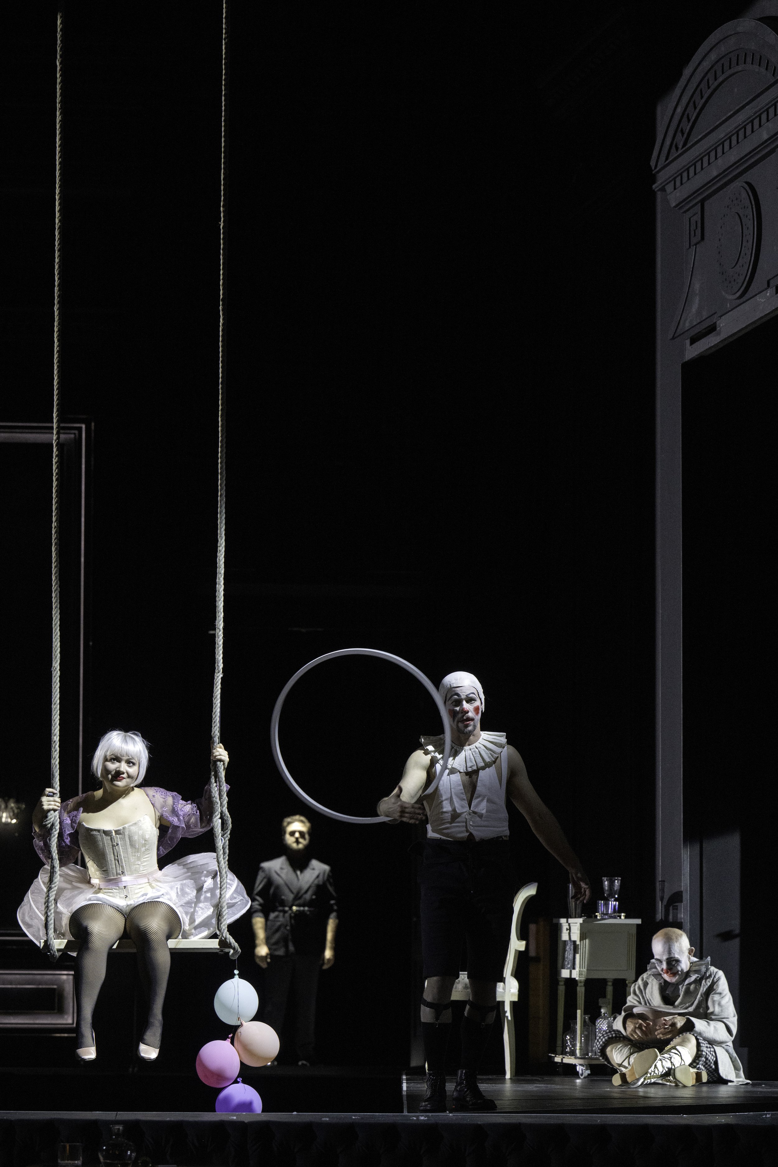 Circus performers on stage with a clown sitting on the floor, a performer on a swing, and a man holding a ring, with balloons hanging from the ceiling.