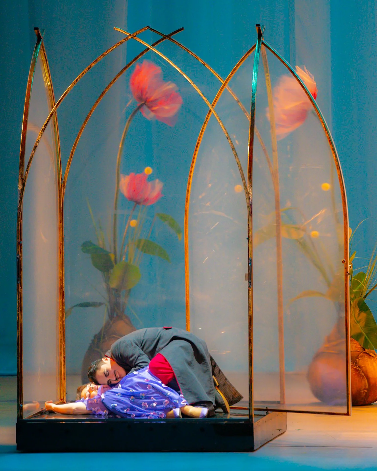 The image shows a woman lying on her side in a glass display case, appearing to be a part of an art installation or performance. The interior background includes painted flowers and a blue backdrop.