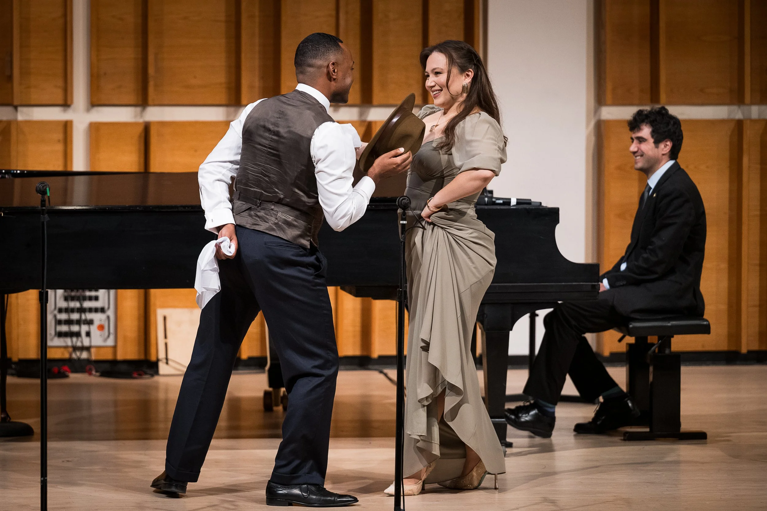 A woman in an elegant dress and high heels smiling at a man in a vest and dress shirt, who is holding a hat. They are on a stage in a music hall, with a pianist sitting at a grand piano in the background. The setting suggests a performance or award c