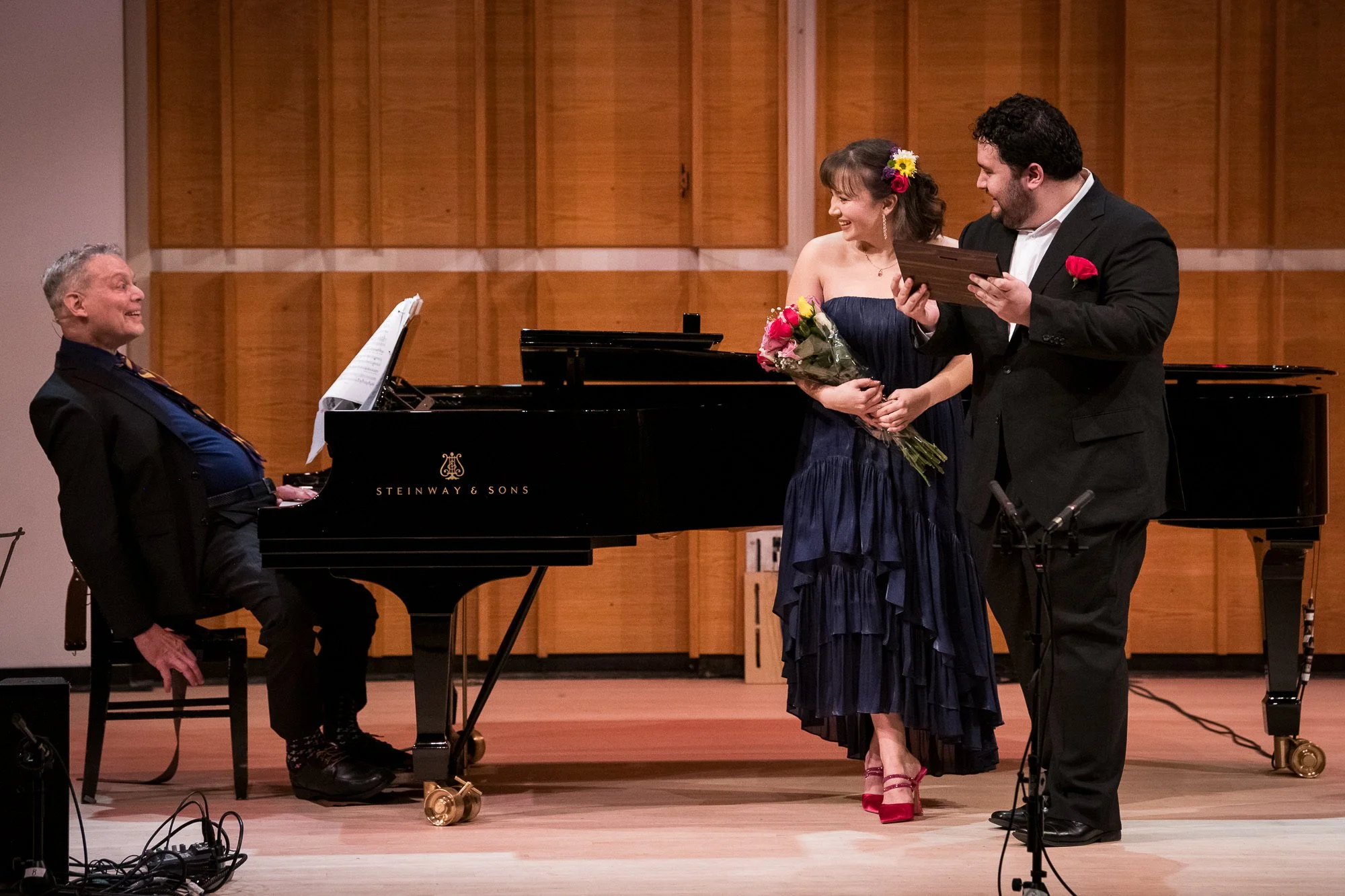 A woman receiving a bouquet of flowers from a man during a musical performance. The seated man plays a grand piano. The background features wooden panel walls.