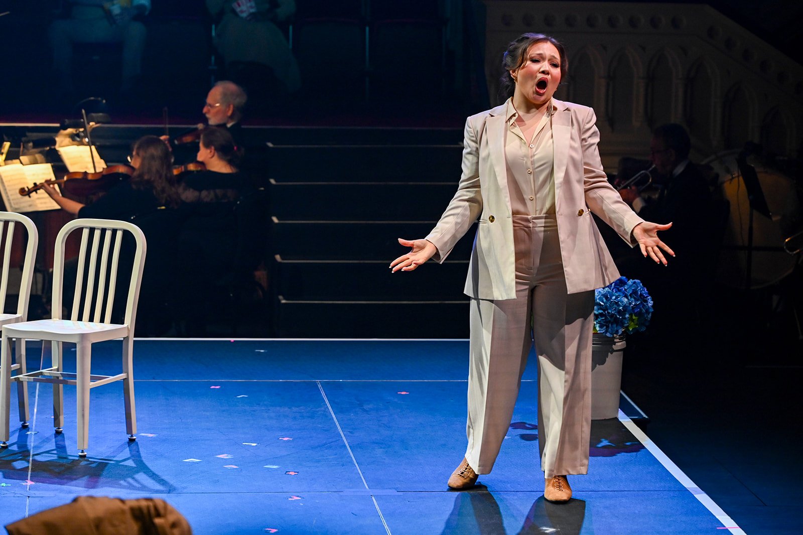 A woman singing on stage with a bouquet of blue flowers beside her, accompanied by an orchestra in the background.