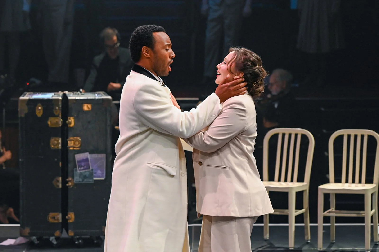 Two actors in white suits are performing on stage, with one actor holding the other's face. In the background, there are chairs and musicians.