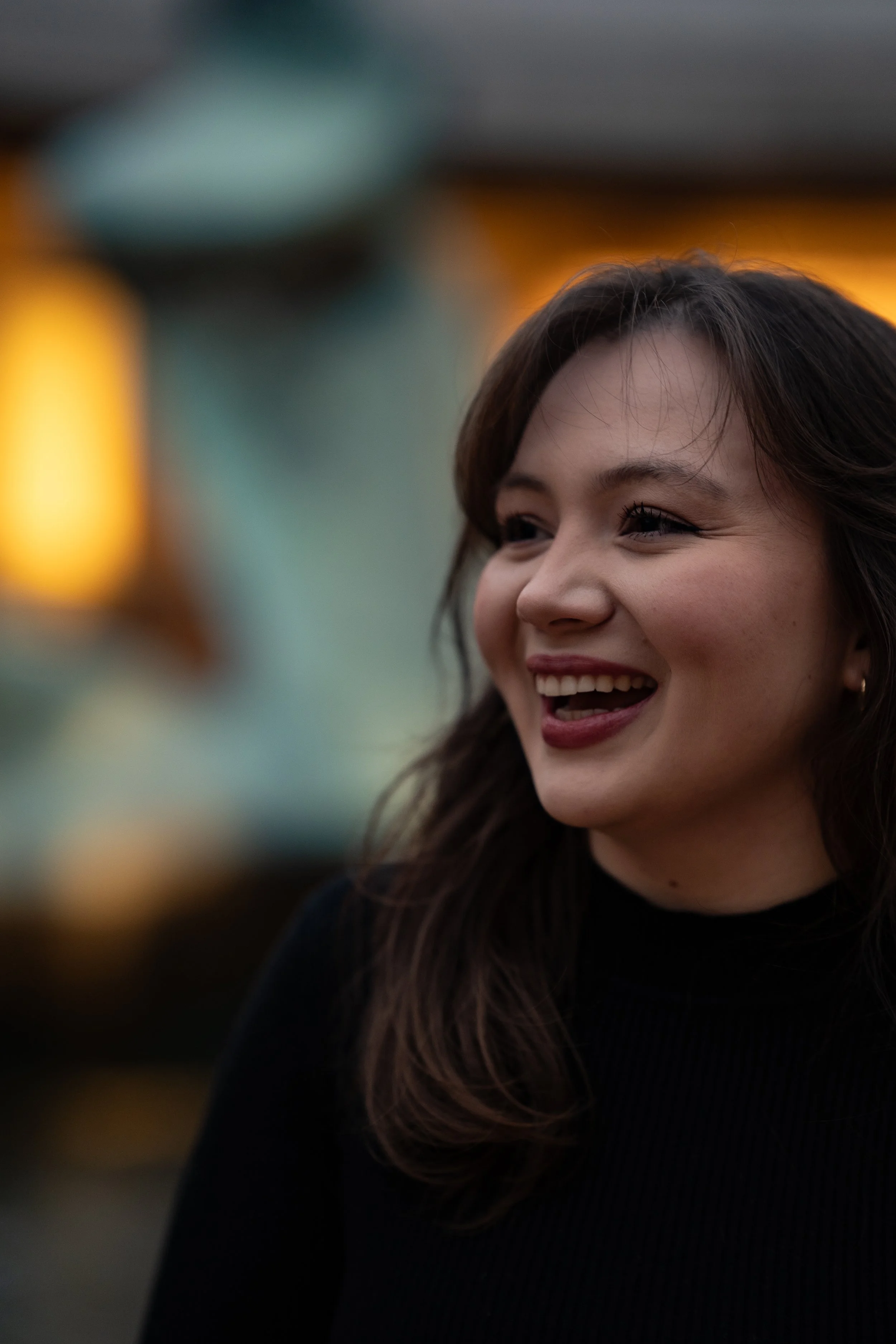 A woman with brown hair smiling and looking slightly to the side, wearing a black top, with blurred yellow and green lights in the background.