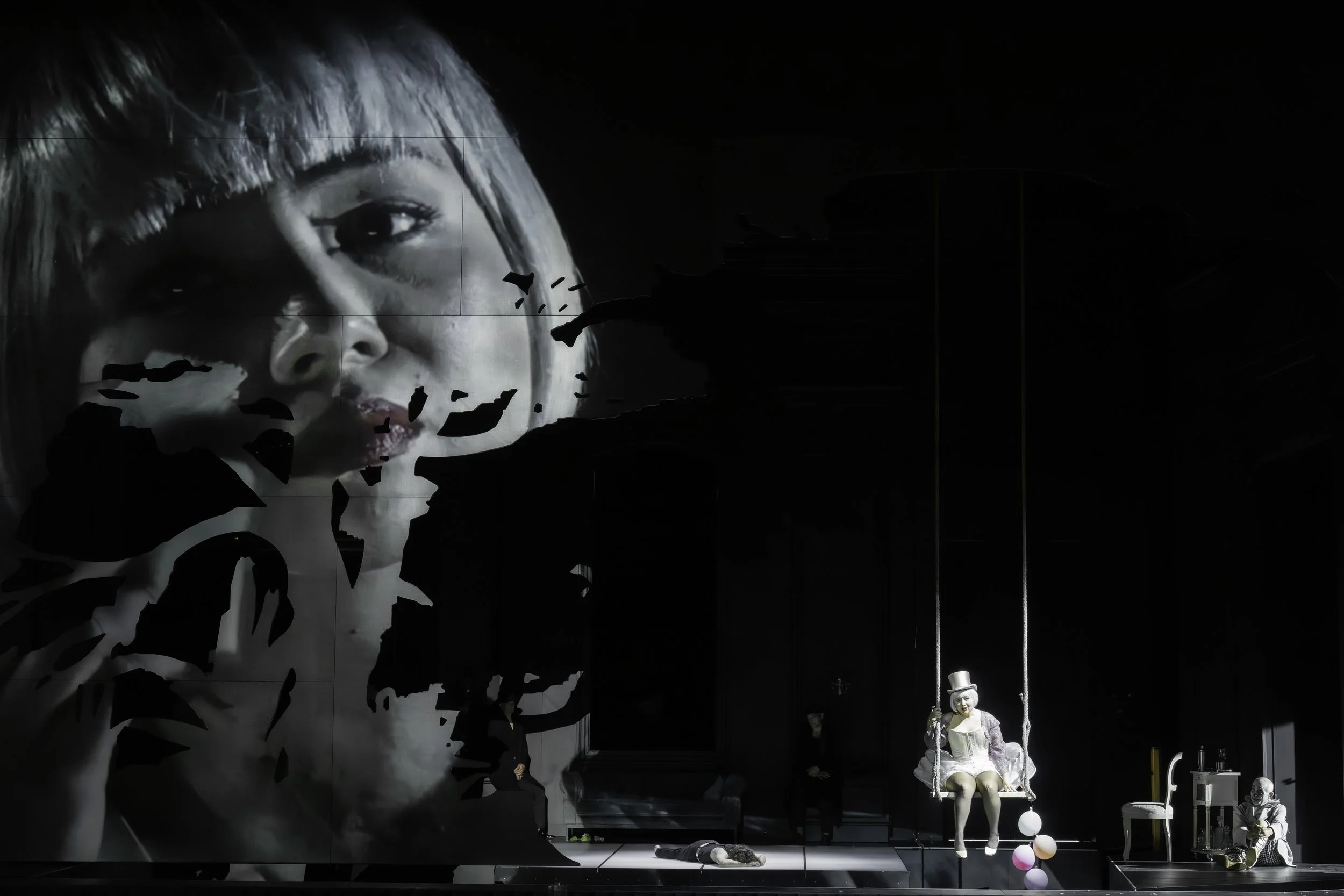 A theatrical scene with a large black and white projection of a child's face on the wall, a mannequin of a girl in a white dress sitting on a swing, and various props like chairs and masks, all set in a darkened room.