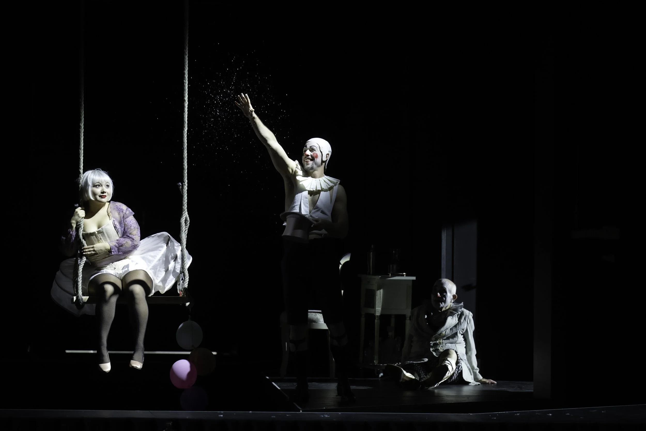 Clowns performing on stage with one sitting on a swing, another standing with a raised arm, and a third kneeling, all with painted faces and theatrical costumes.