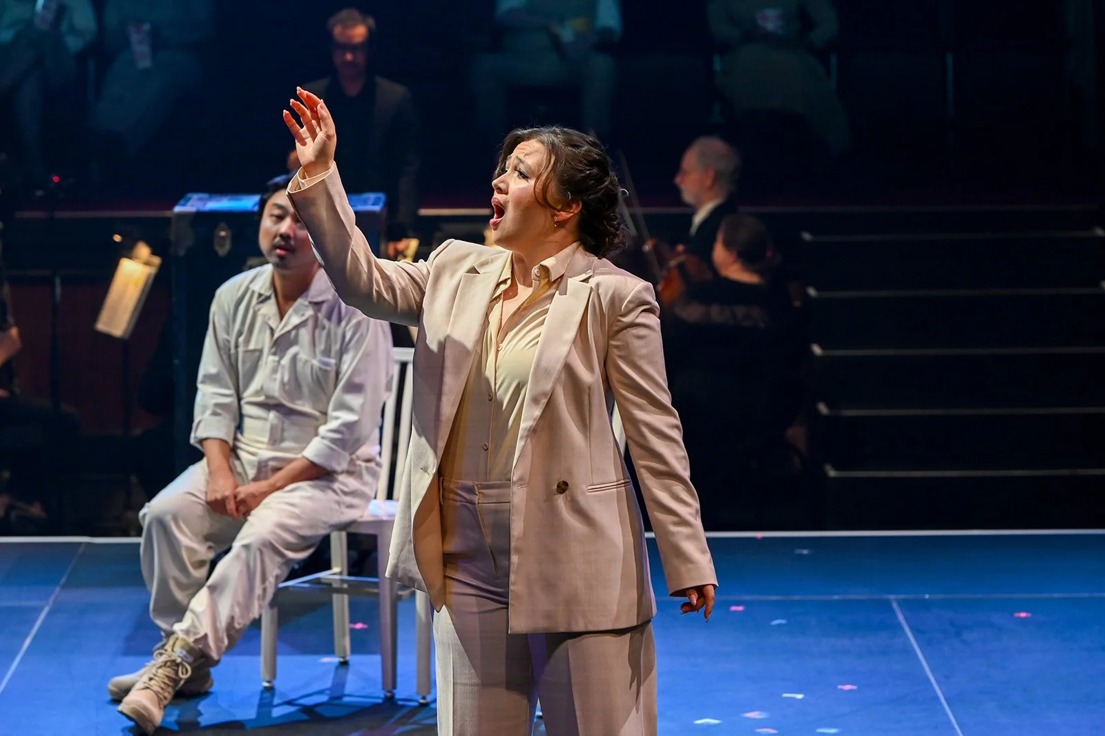 A woman performing on stage with her hand raised, wearing a cream-colored suit, with a man sitting on a chair in the background and musicians playing instruments behind them.
