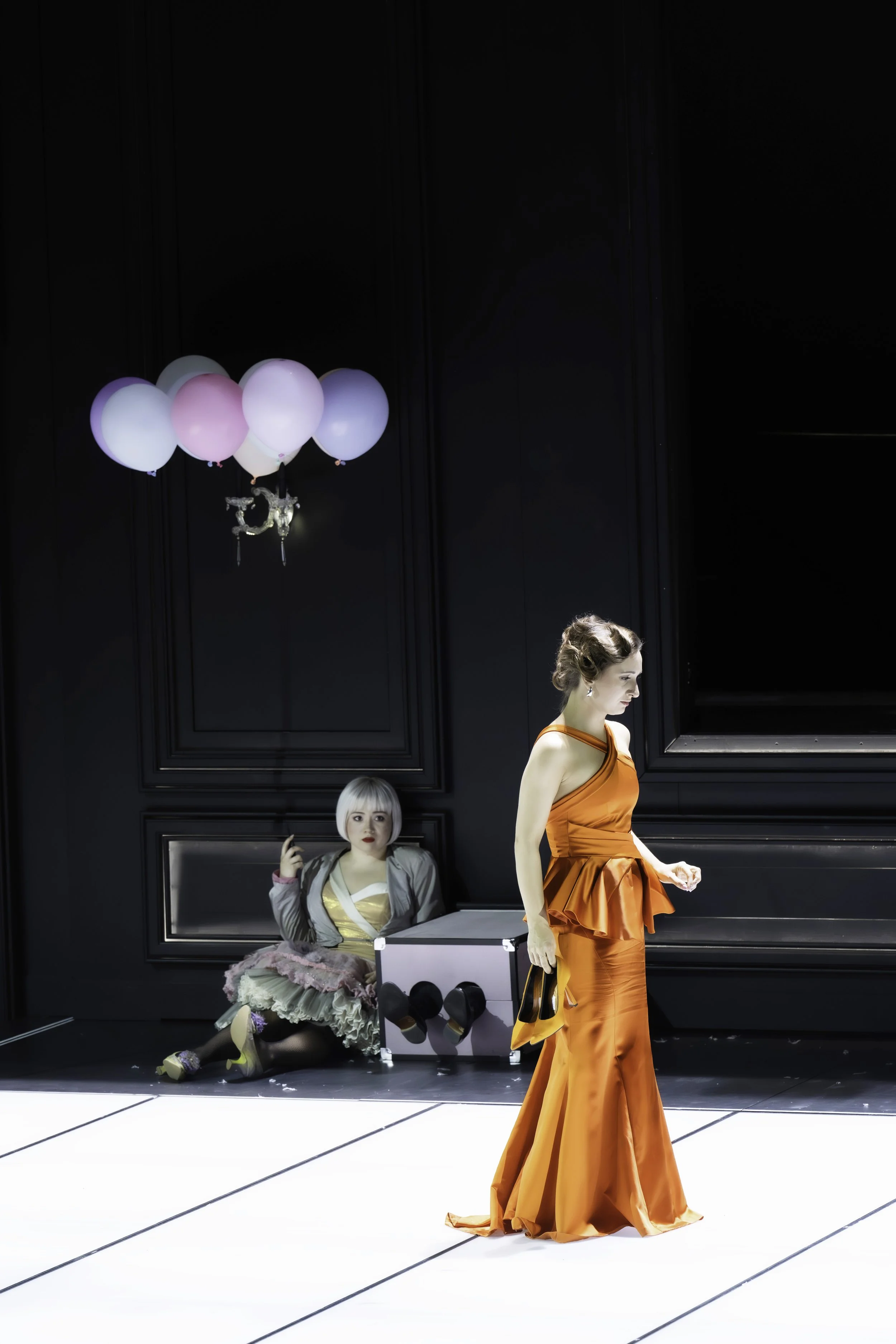 Two women in a fashion runway scene. One woman in an orange dress is walking, while the other woman in a floral and beige dress is sitting on the floor and talking on a cellphone. There are pastel-colored balloons attached to two helium balloons in t