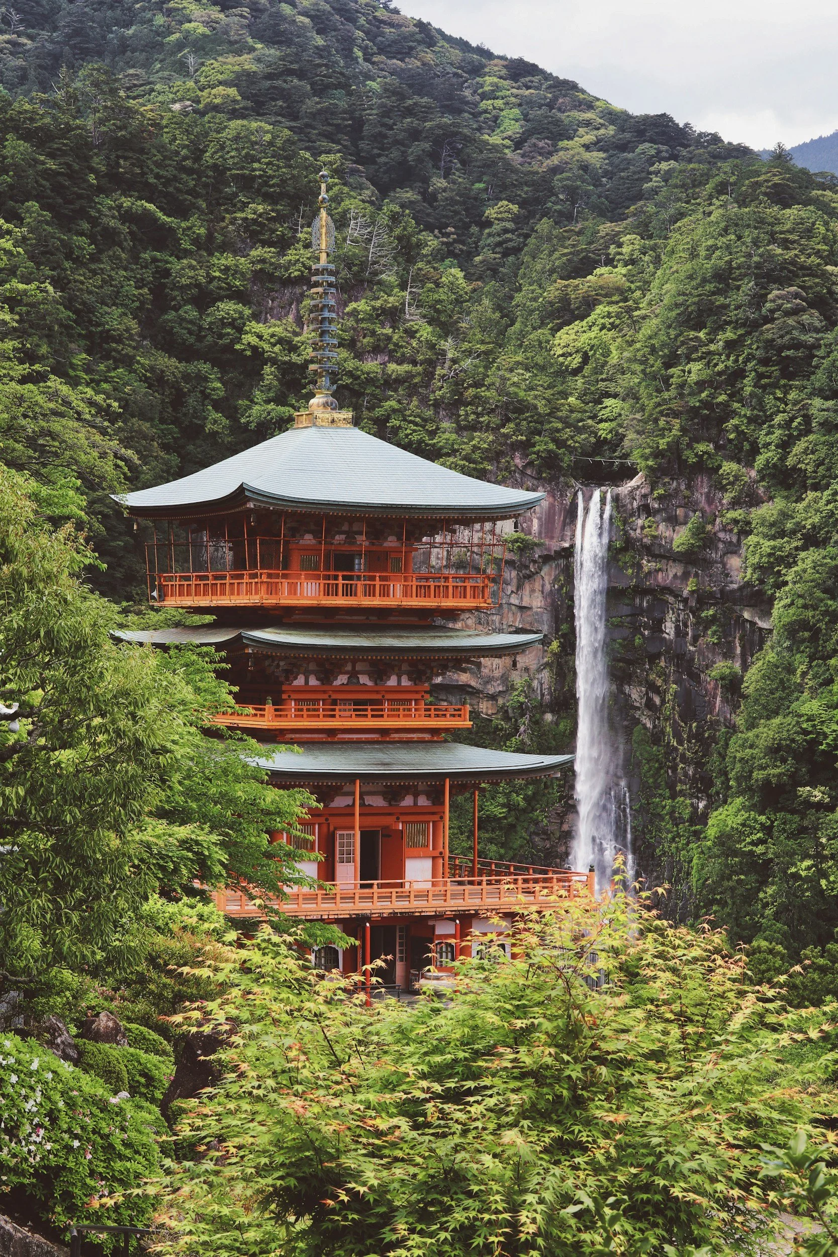 A traditional Japanese pagoda with multiple tiers surrounded by lush green trees, with a waterfall cascading down the forested mountain in the background.