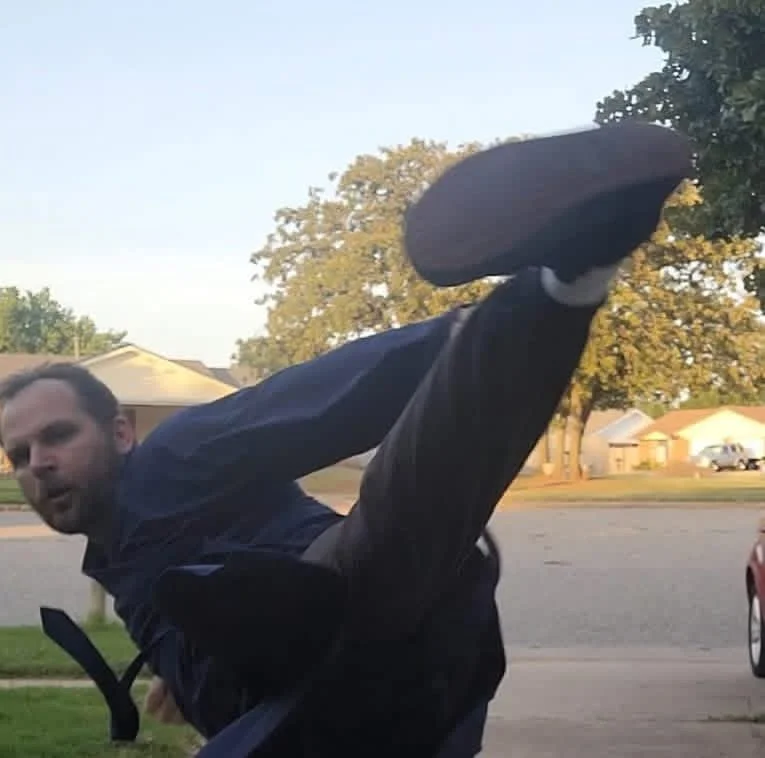 Man in dark clothing executing a high kick outdoors during daytime, with a suburban neighborhood and trees in the background.