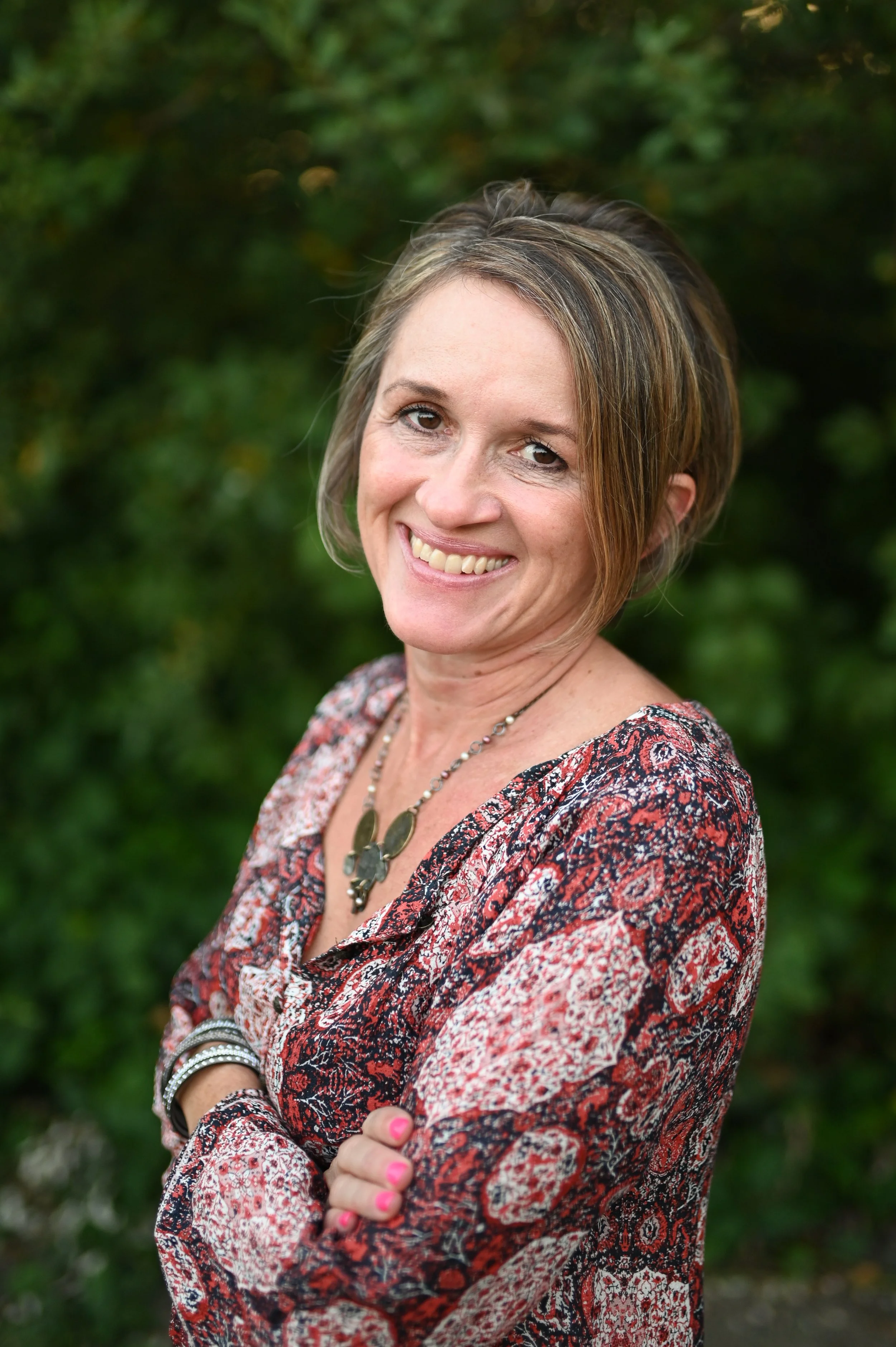 A woman with short hair and a smile, wearing a patterned blouse and layered necklaces, standing outdoors with blurred greenery in the background.