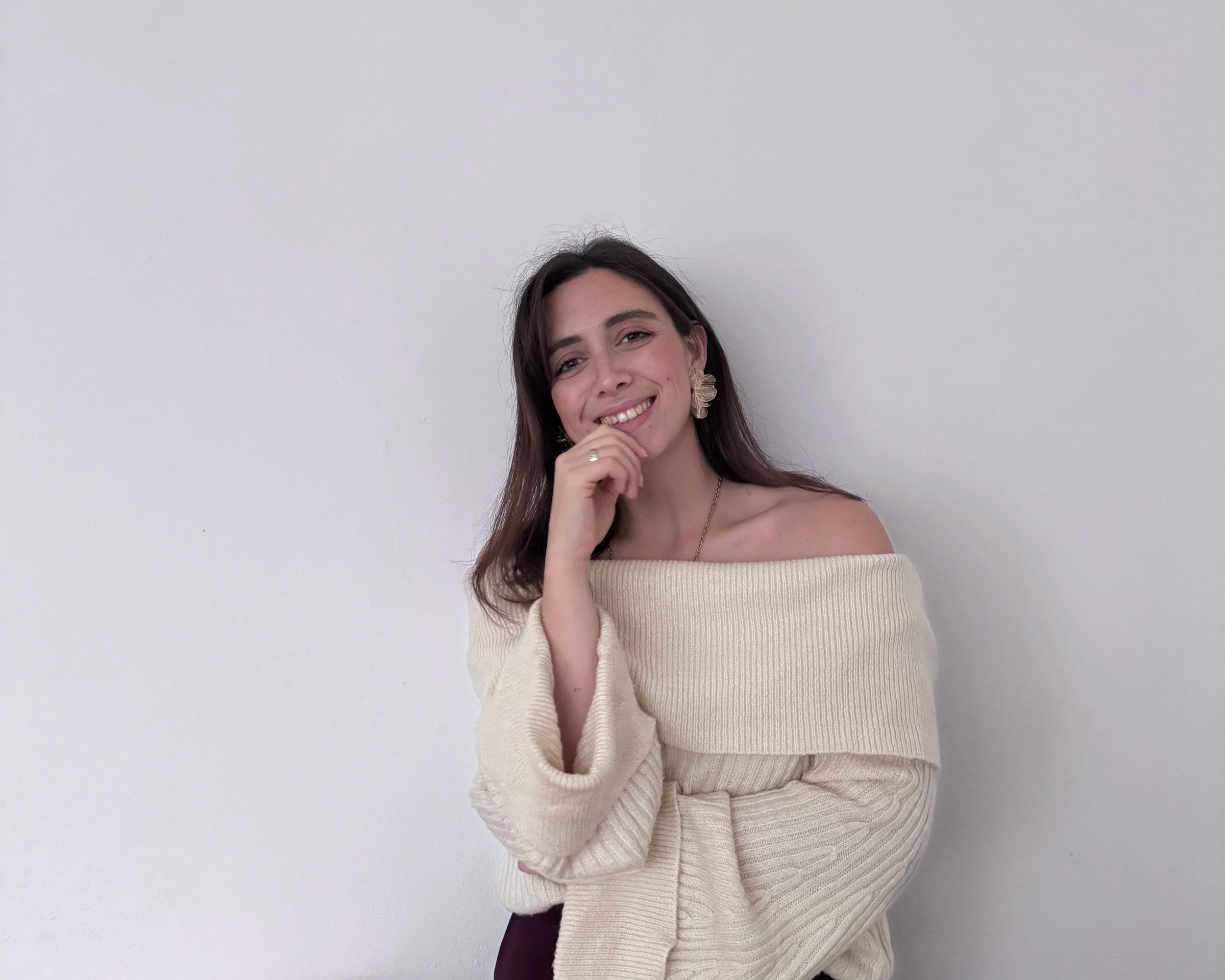 Eine junge Frau mit braunen Haaren, die ein off-the-Shoulder cremefarbenes Strickpullover trägt, lächelt und die Hand an das Kinn legt, vor einer weißen Wand.