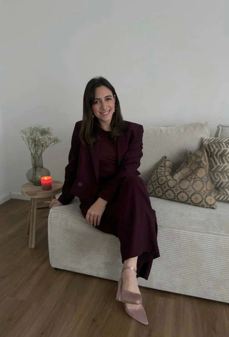 Eine junge Frau sitzt auf einer sofaschicht mit dunkler Hose und Blazer, lächelt und hat dunkle, glatte Haare. Im Hintergrund steht ein kleiner Beistelltisch mit einer Vase mit weißen Blumen und einer roten Kerze.