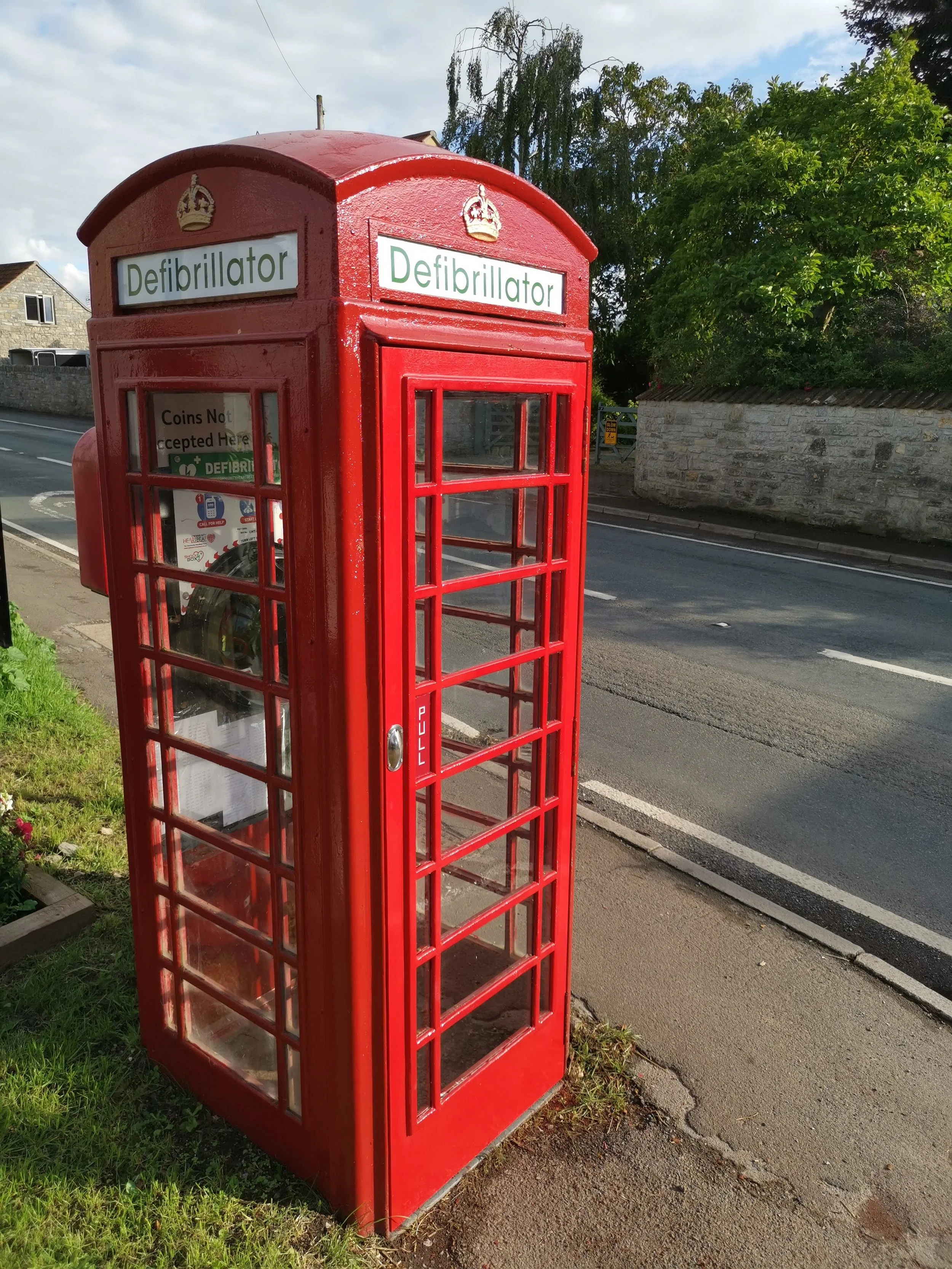 Traditional red British telephone booth with a sign that says 'Defibrillator' at the top, located on a sidewalk next to a road with houses and trees in the background.