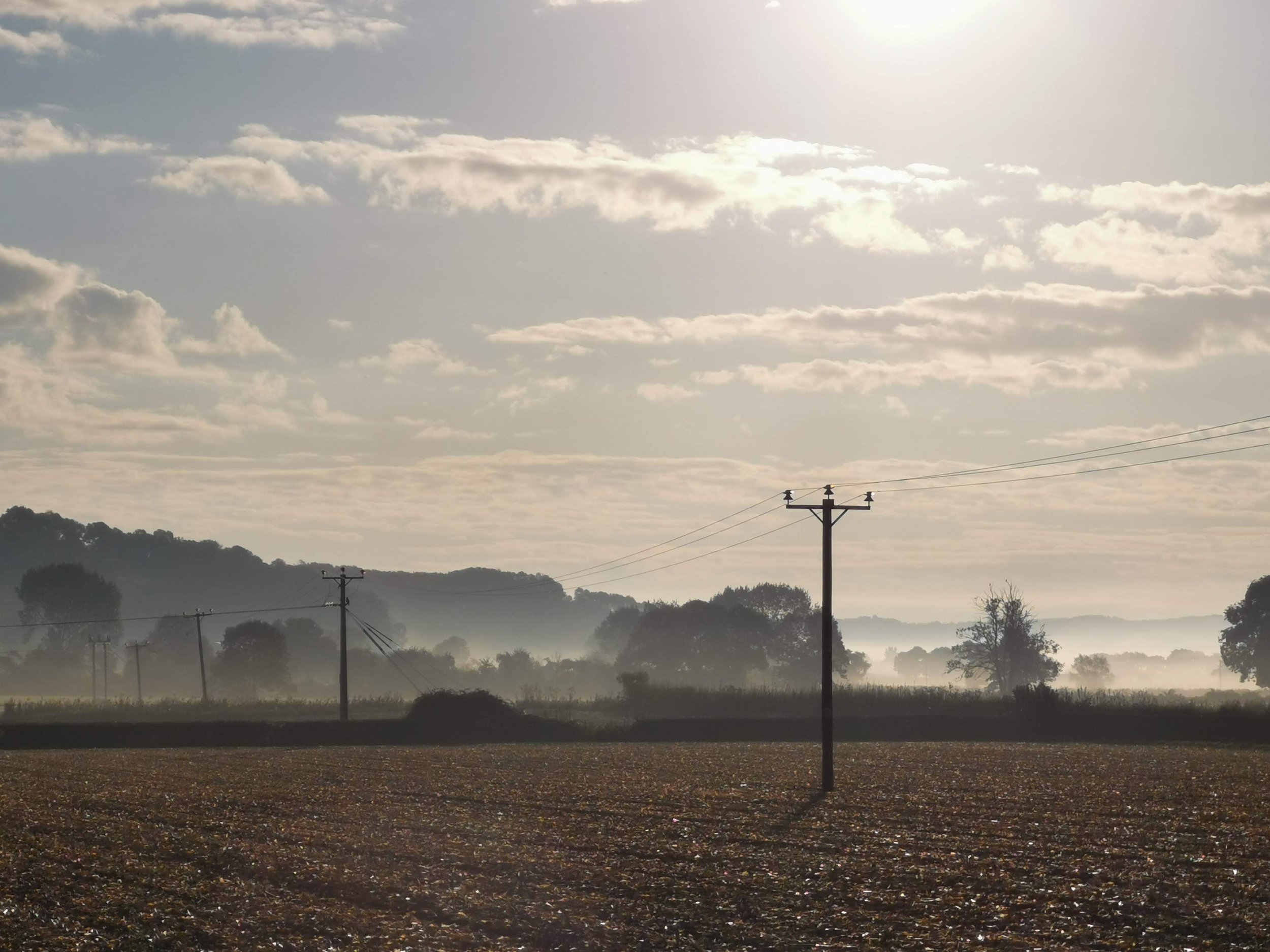 A rural landscape with utility poles running through a plowed field, trees in the distance, a misty horizon, and a sky with scattered clouds and bright sunlight.
