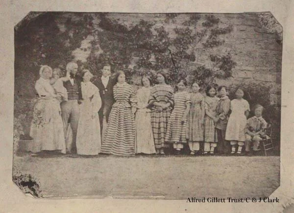Black and white photo of a group of children and young adults standing outdoors in front of trees, some in striped dresses, with one seated child on a chair at the right end.