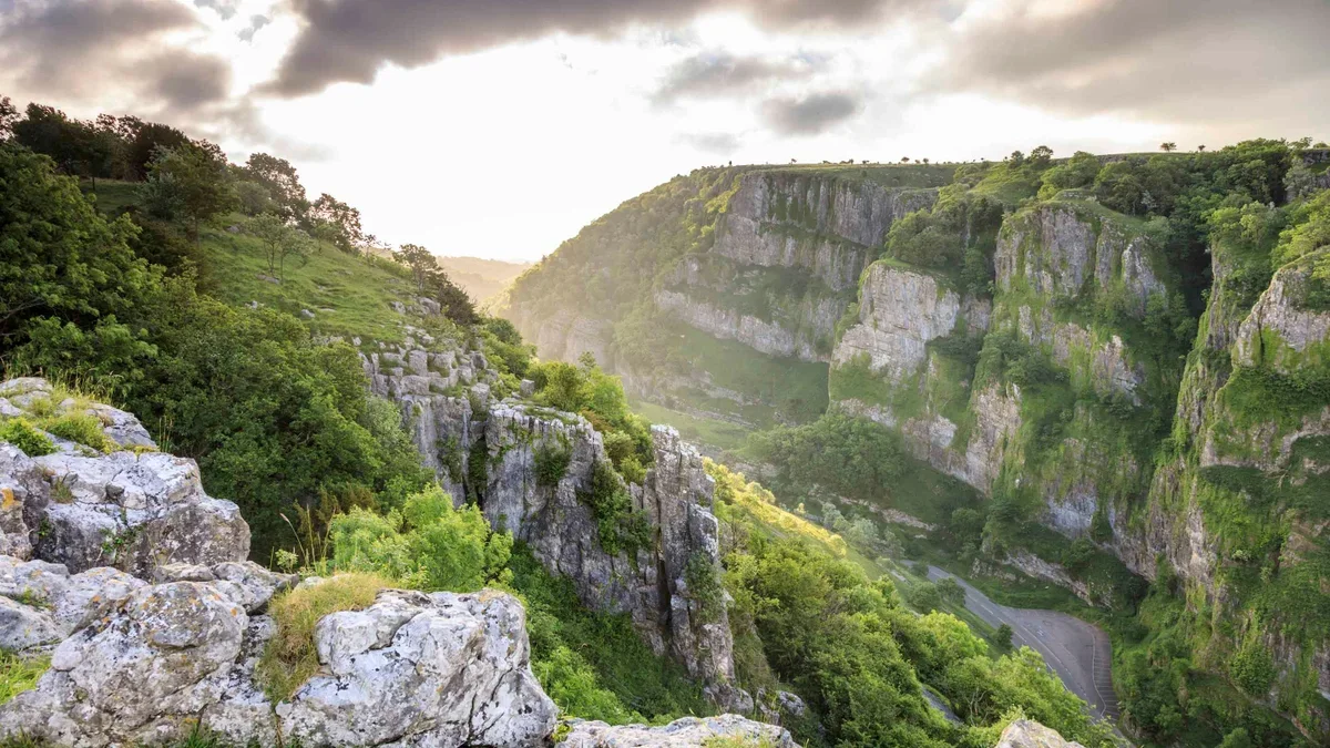 A scenic view of a lush green valley with steep rocky cliffs on either side, and a road or trail winding through the bottom of the valley, under a cloudy sky with the sun shining through.