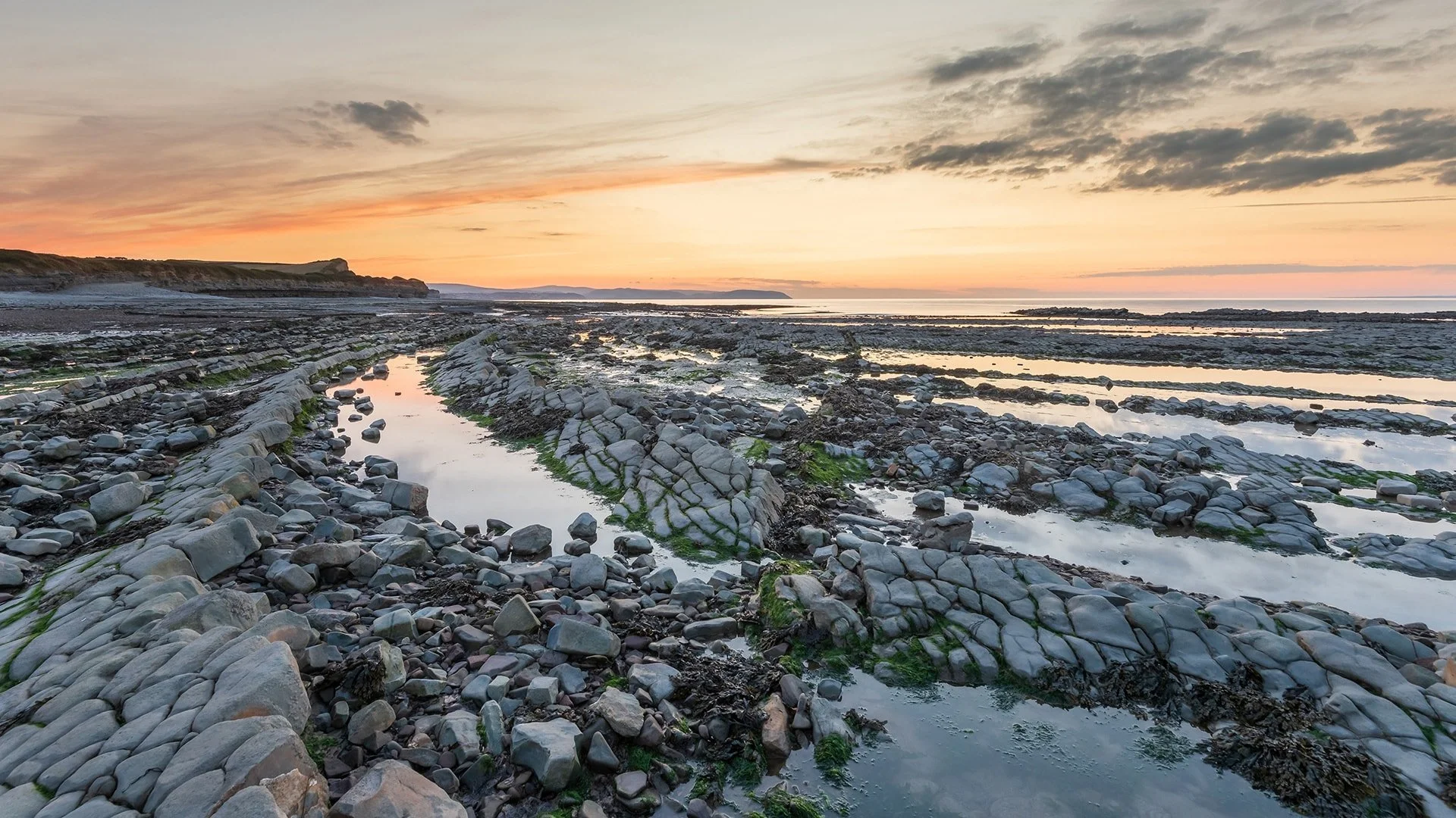 Sunset over a rocky coastal shoreline with tide pools, scattered seaweed, and distant cliffs.