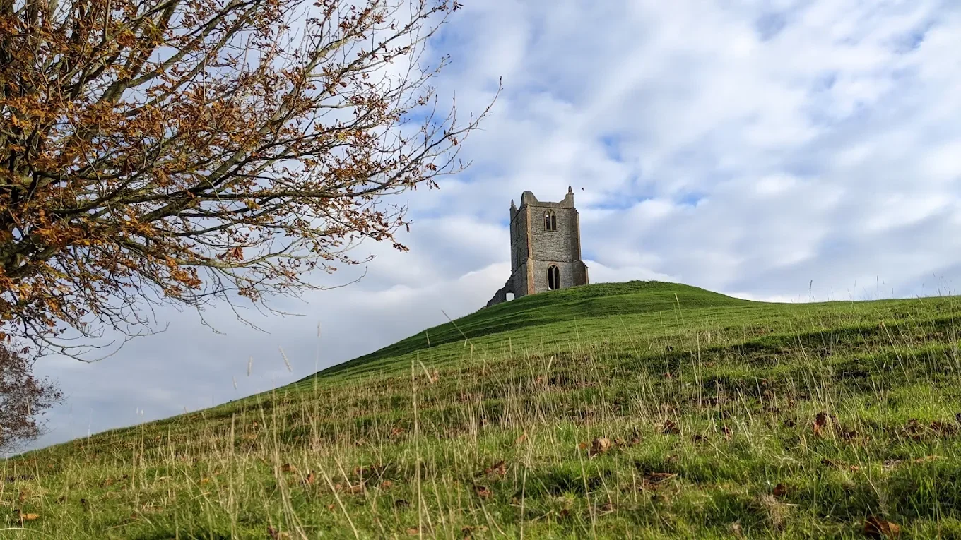 An old stone tower built on top of a grassy hill, with a tree with sparse leaves on the left and a partly cloudy sky in the background.