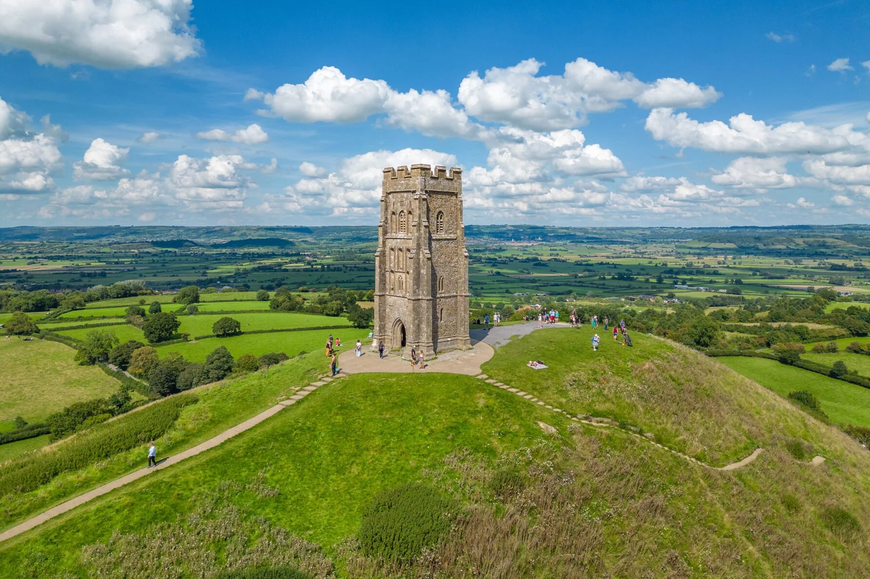 A stone tower on a grassy hilltop with a dirt path leading to it, surrounded by green fields and a bright blue sky with scattered white clouds.