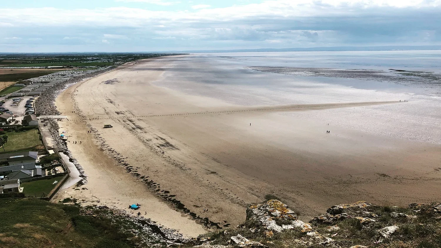 A wide view of a sandy beach with some houses and parked cars on the left side. There is a line of rocks at the foreground and a stone breakwater extending into the water. The sky is partly cloudy with patches of blue.