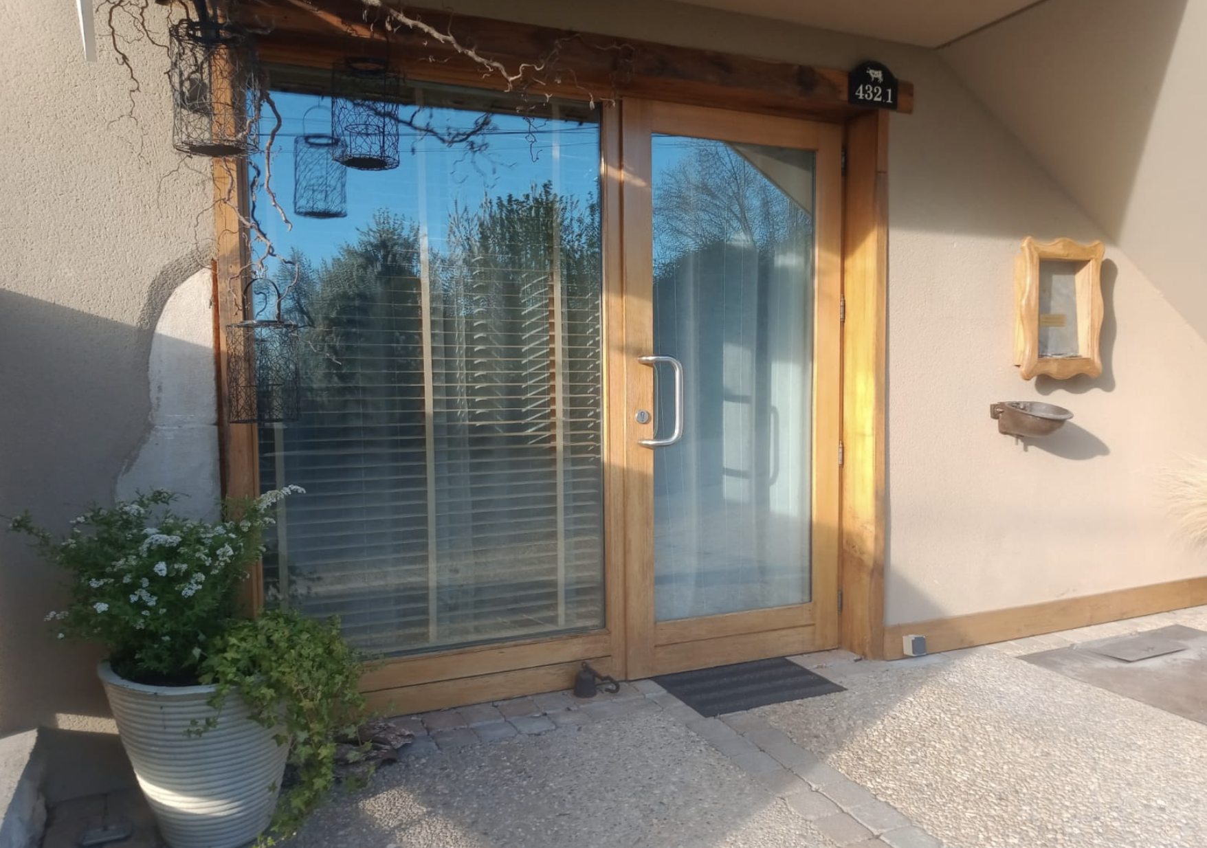 A wooden framed glass door with a silver handle, flanked by a beige wall with wall-mounted decorations including a small picture frame, a small ceramic dish, and house number 432.1. To the left of the door, outdoor plants in a white pot and decorative black wire baskets hanging above. The area appears to be a patio or entranceway with sunlight casting shadows.