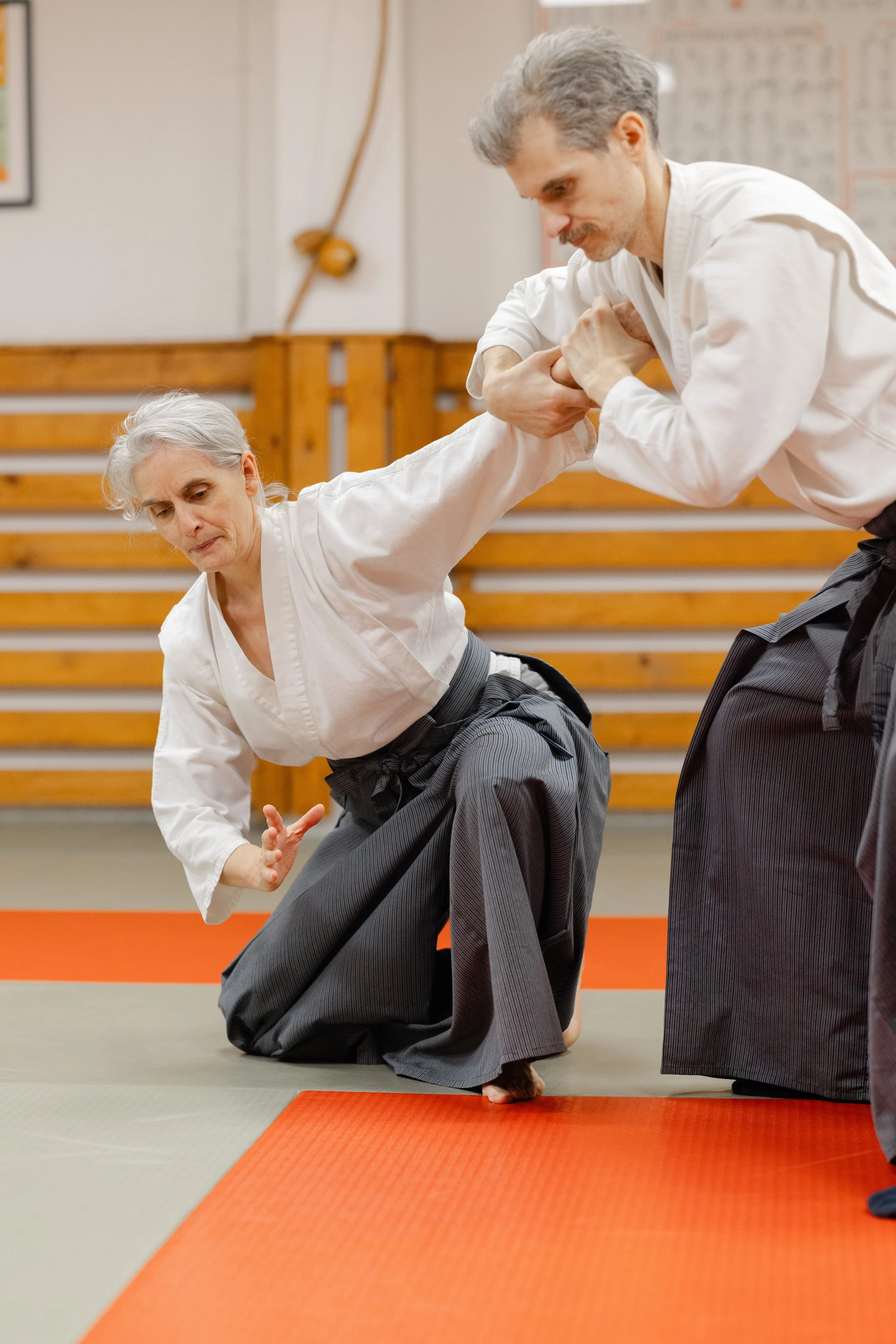 A martial arts instructor demonstrates a technique with an elderly woman on a padded training mat in a dojo. Both are dressed in traditional white uniforms with gray gi pants, and the woman is in a kneeling position while the instructor holds her arm during the demonstration.