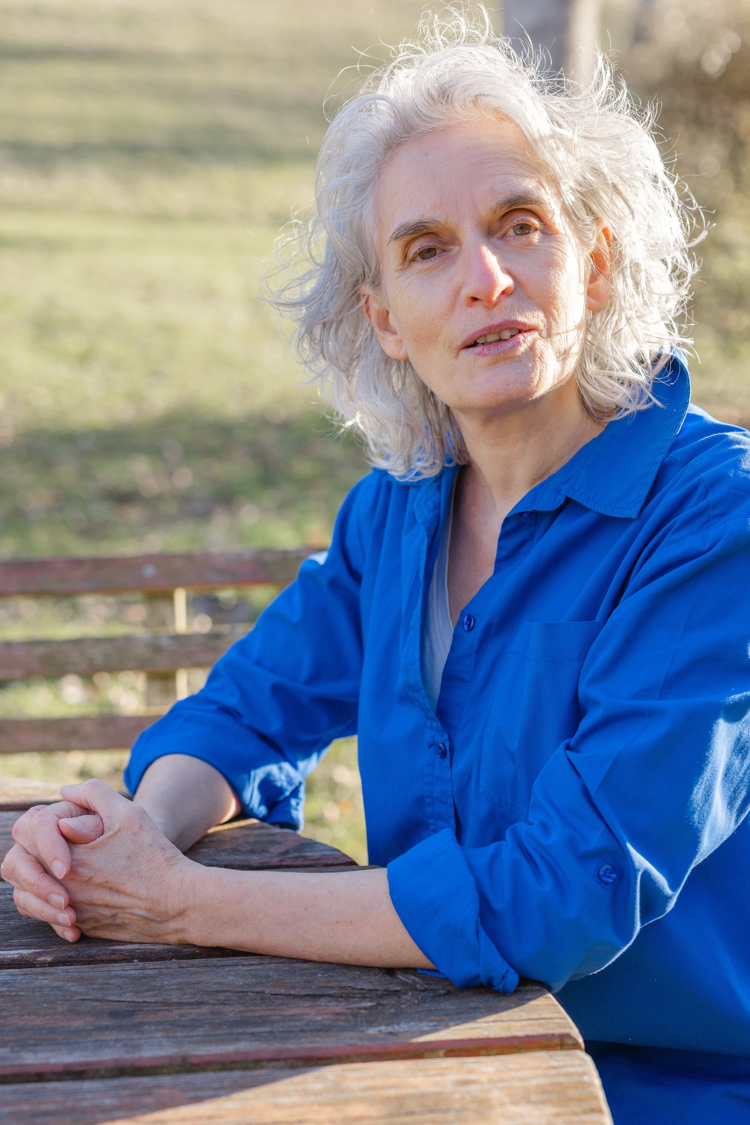 Older woman with gray hair sitting outdoors at a park bench, wearing a blue shirt and looking at the camera.