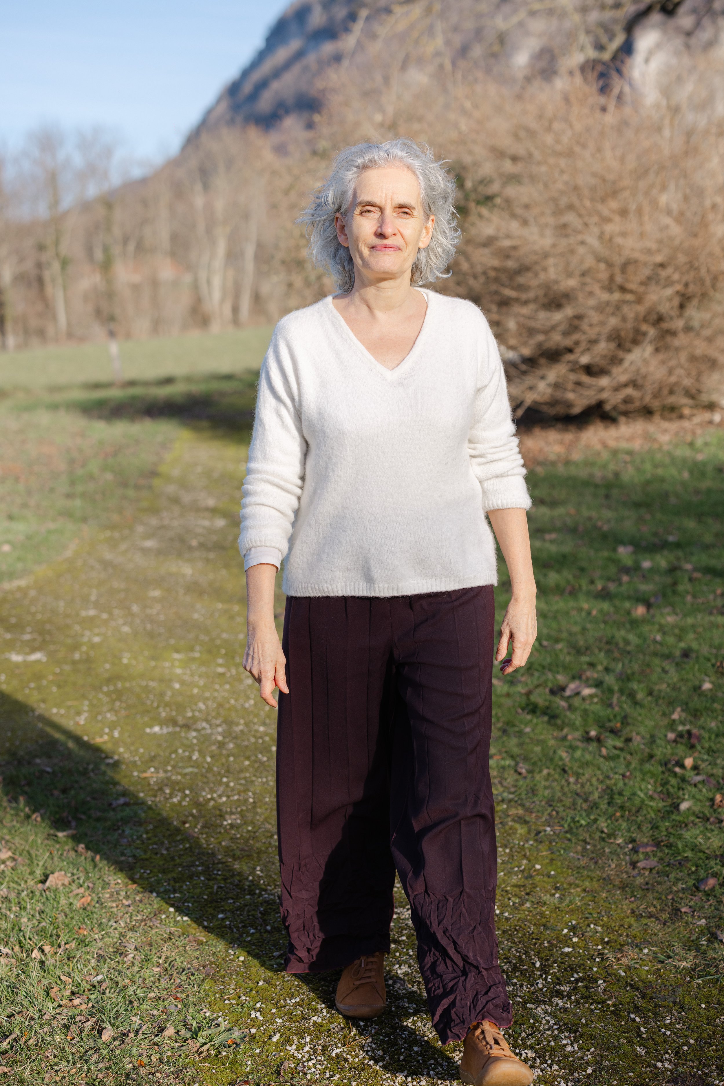Older woman with gray hair walking outdoors on a grassy and gravel path, wearing a white sweater, dark pants, and tan boots, with trees and a mountain in the background.