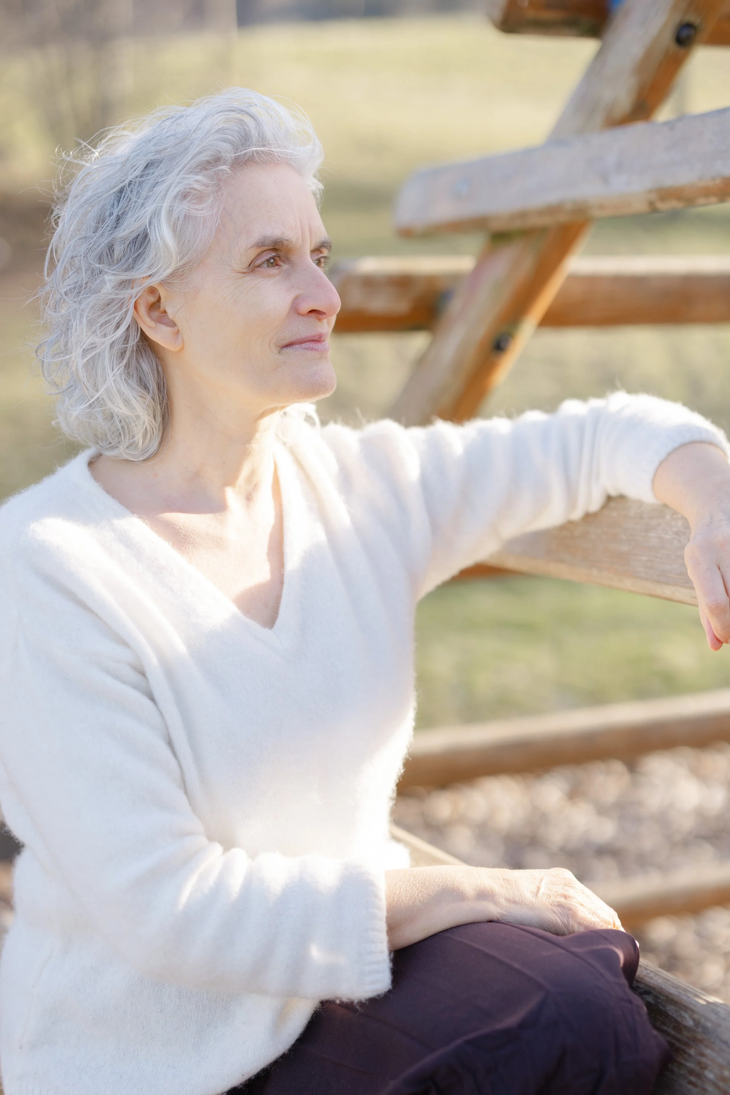 An elderly woman with gray hair sitting on a wooden bench outdoors, looking into the distance with a calm expression, wearing a white sweater and dark pants.