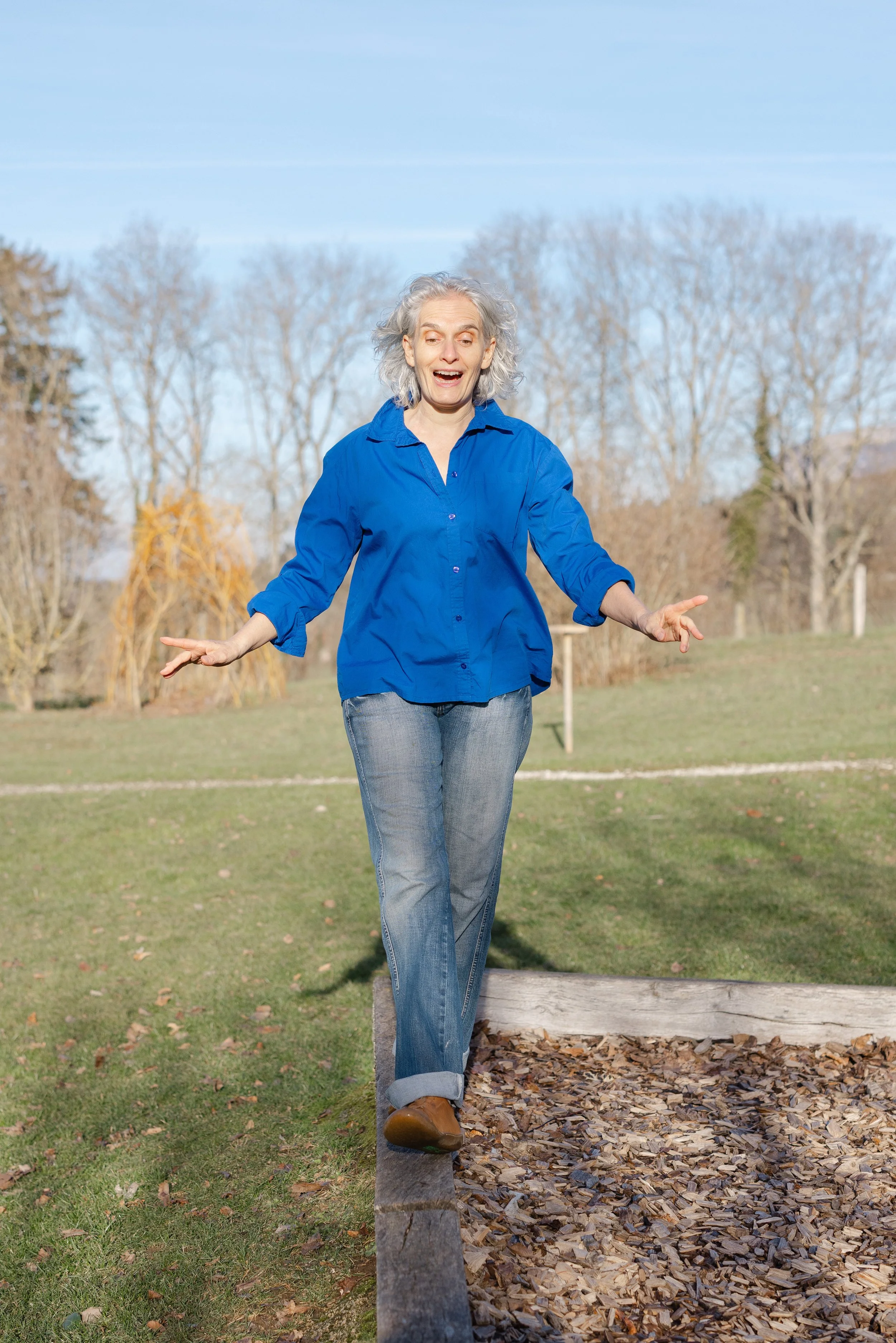 An elderly woman with white curly hair, wearing a bright blue shirt and jeans, balancing on a wooden beam in an outdoor park during daytime, with leafless trees and a clear blue sky in the background.