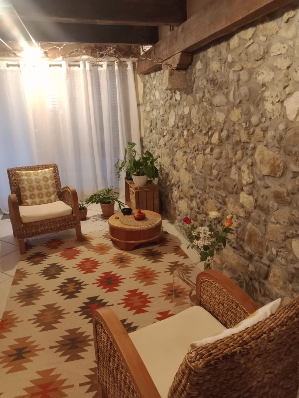 A cozy indoor space with a stone wall, white curtains, wicker chairs with cream cushions, and a geometric patterned rug. There are potted plants and a round wooden coffee table with decorative items.