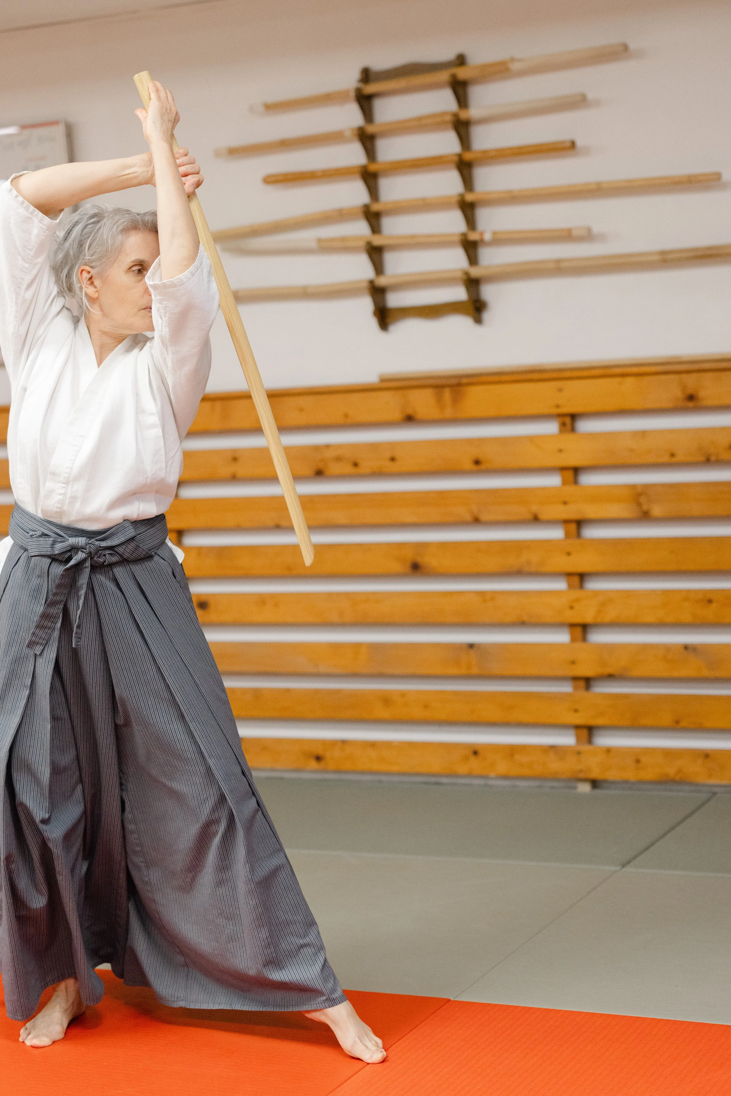 A person practicing martial arts or possibly performing a kata in a dojo, wearing traditional clothing, standing on a red martial arts mat with wooden wall bars in the background.