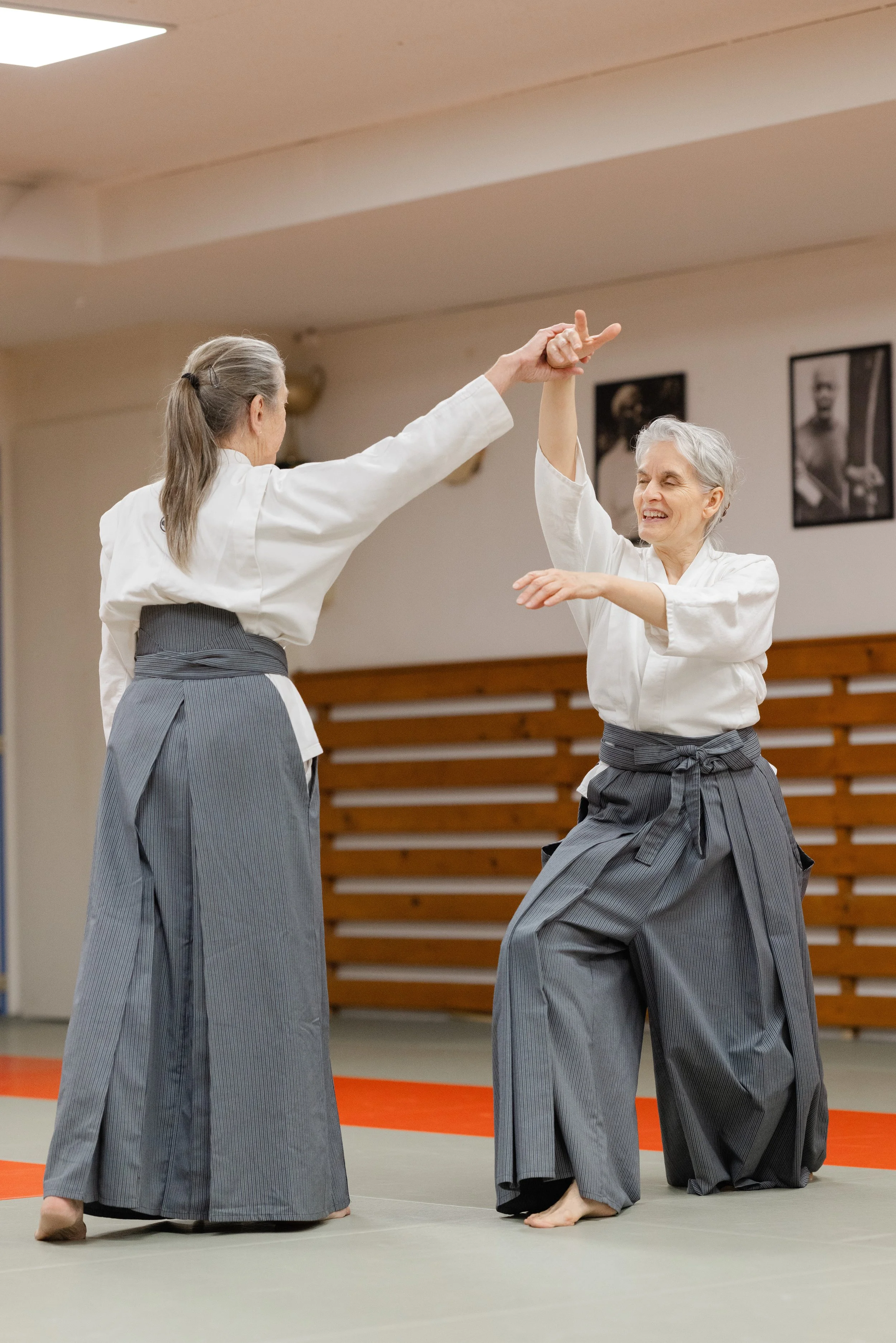 Two women practicing Aikido in a dojo, one kneeling with a sword, the other standing, both in traditional martial arts uniforms, with framed photographs on the wall behind them.