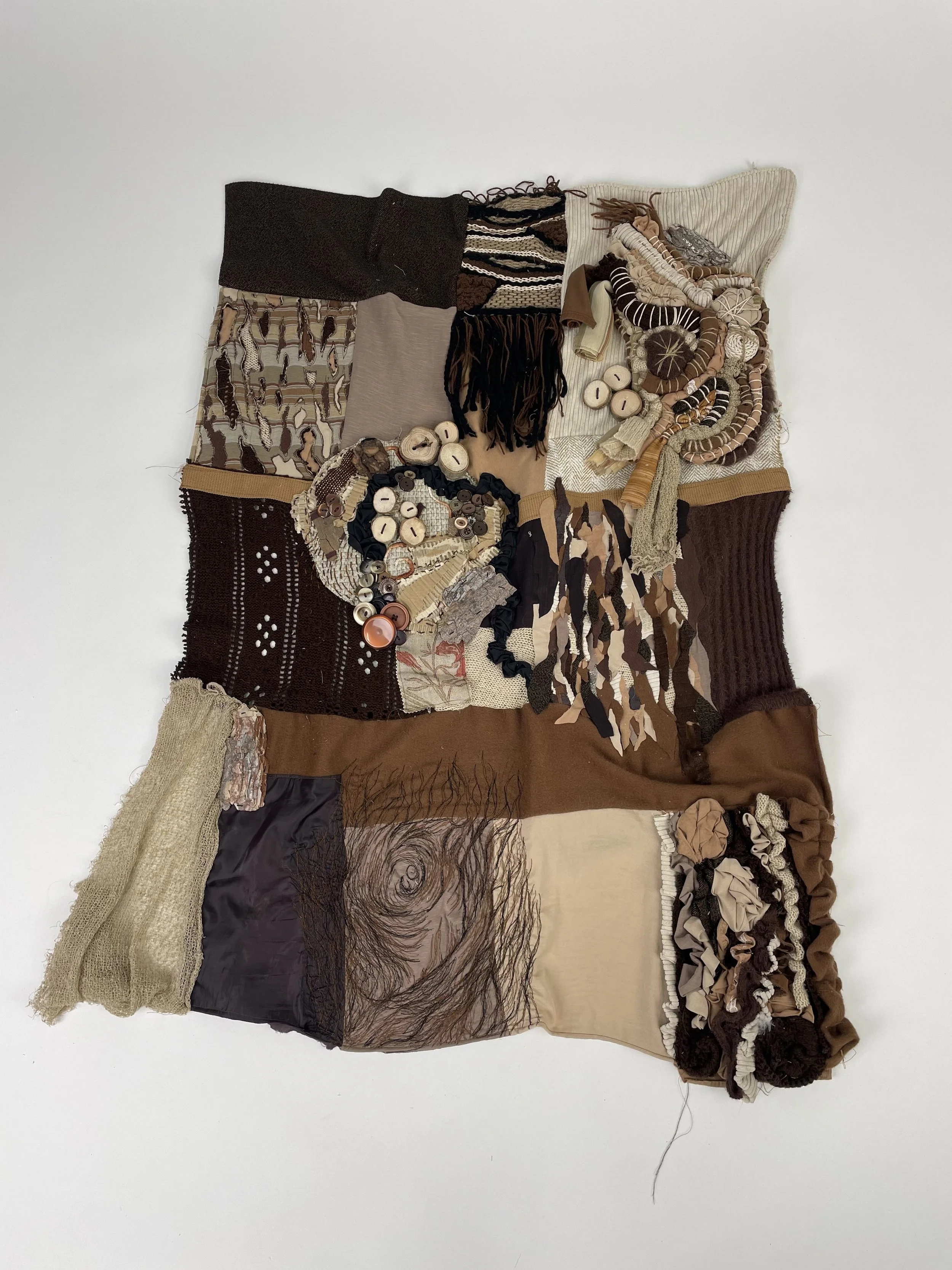A mixed-media artwork made from various fabric pieces, buttons, and other materials assembled on a white background, resembling a collage or textile art piece.