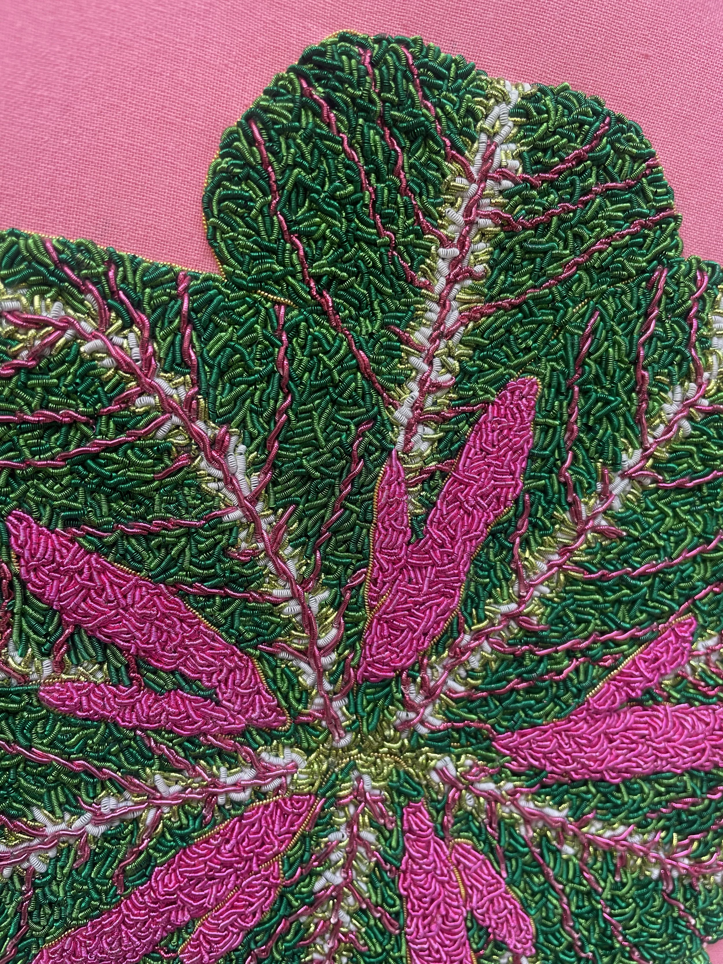 Close-up of detailed bead embroidery of a pink flower with green leaves on pink fabric background.