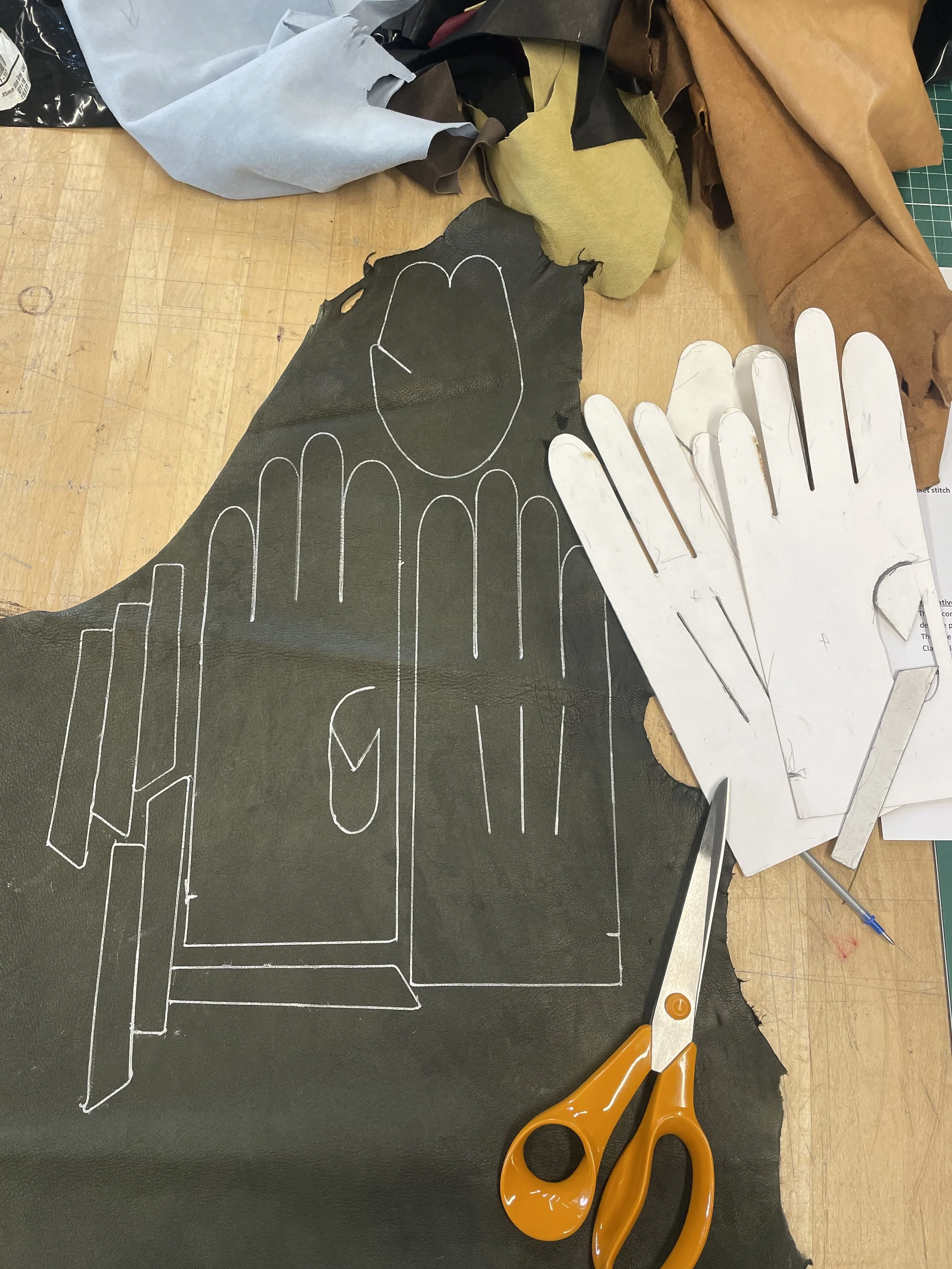 A workspace with leather pieces, pattern templates, orange scissors, and a utility knife on a black surface. The leather pieces have white chalk outlines for cutting.