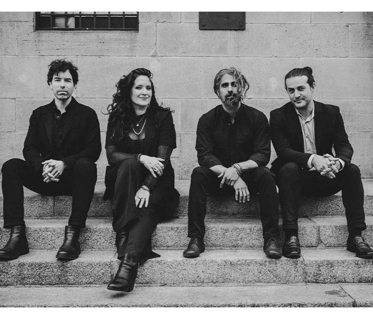 Four people sitting on concrete steps against a brick wall, dressed in dark clothing, with two men and two women, a black and white photo.