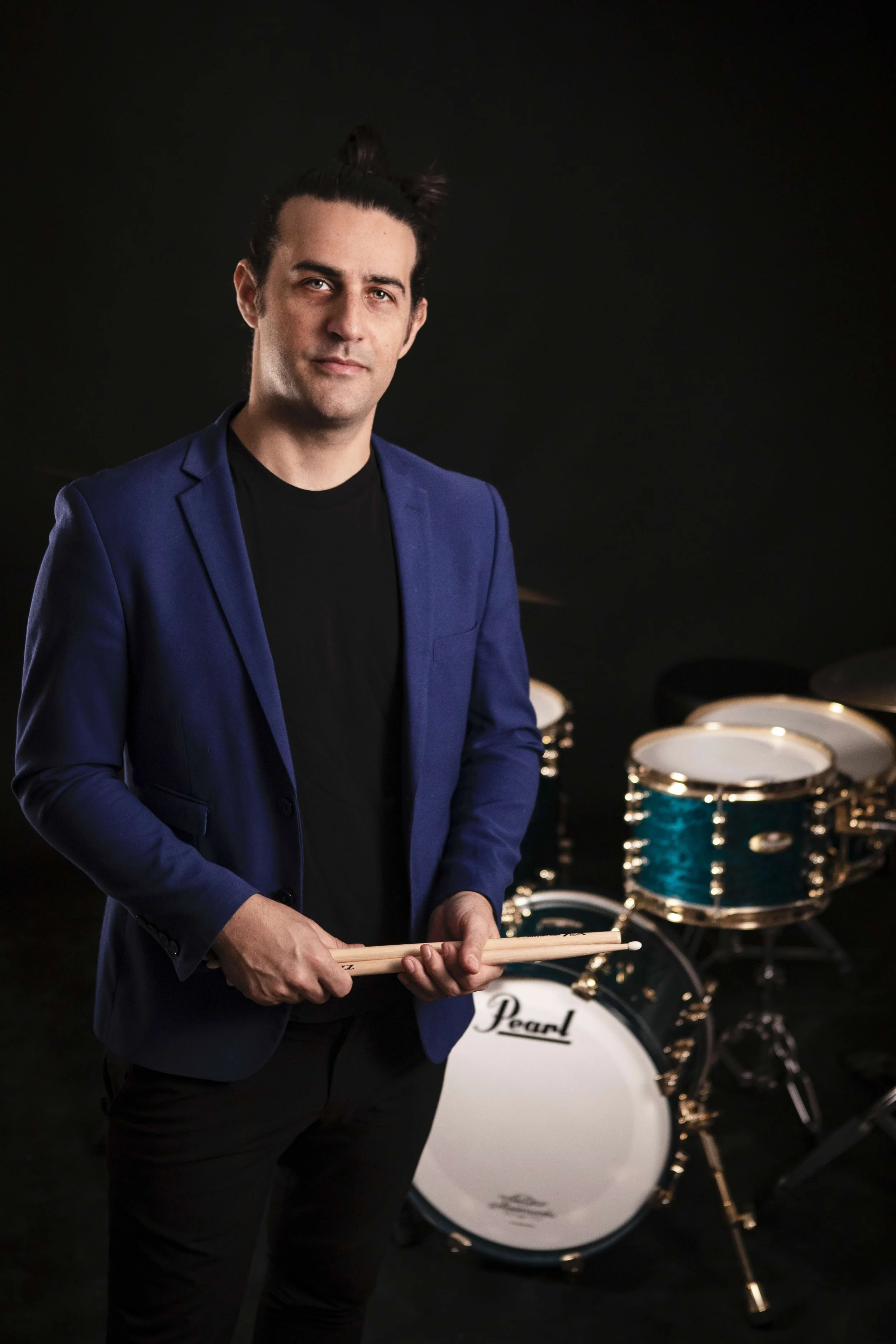 Drummer Dan Pugach in a blue blazer holding drumsticks stands in front of a blue drum set against a black background.