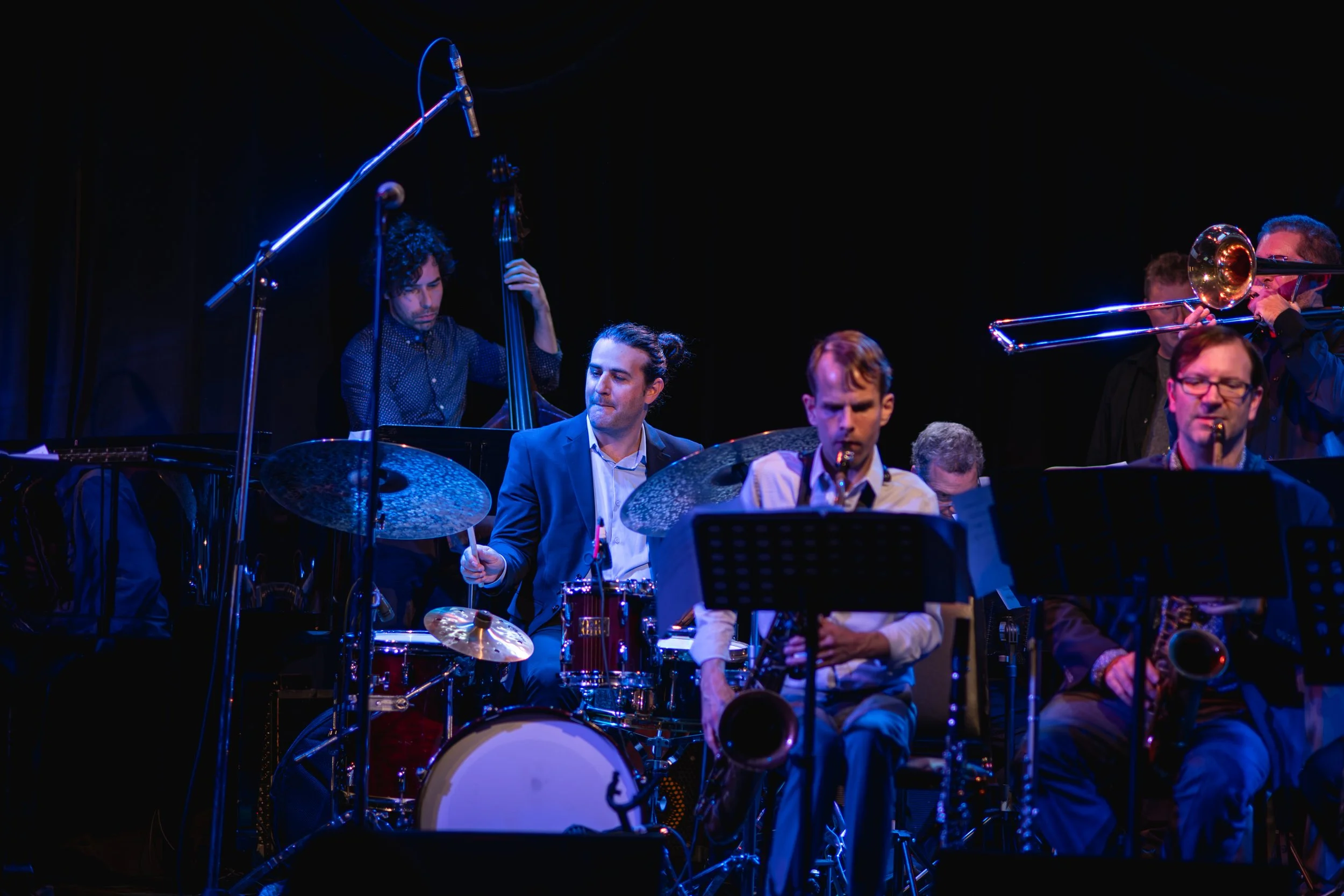 Jazz band performing on stage with musicians playing drums, double bass, trombones, and saxophones under stage lighting.
